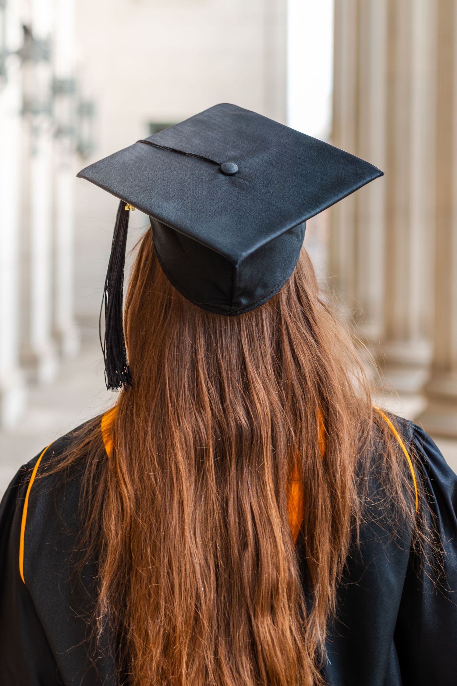Graduate in black cap and gown with long red hair stands before classical columns at graduation ceremony.