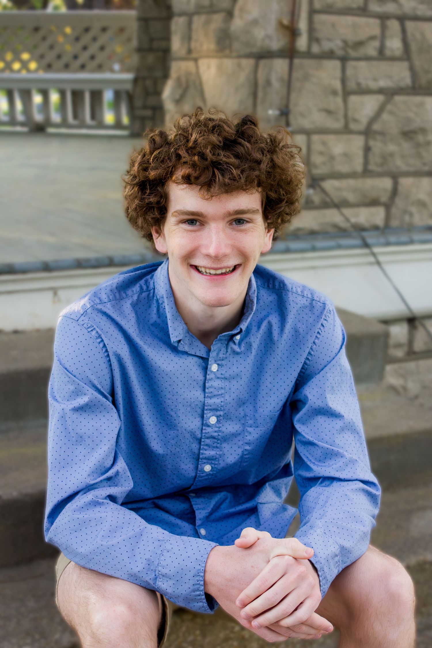 Someone in a light blue polka dot button-down shirt with curly hair sitting outdoors near stone architecture.