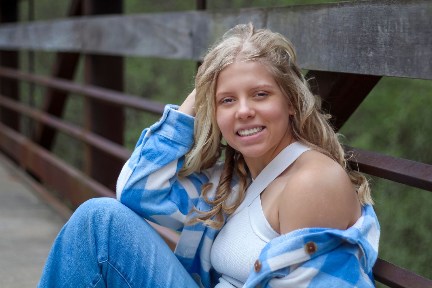 Person in casual blue jacket and white top sits on wooden bridge railing with natural background.
