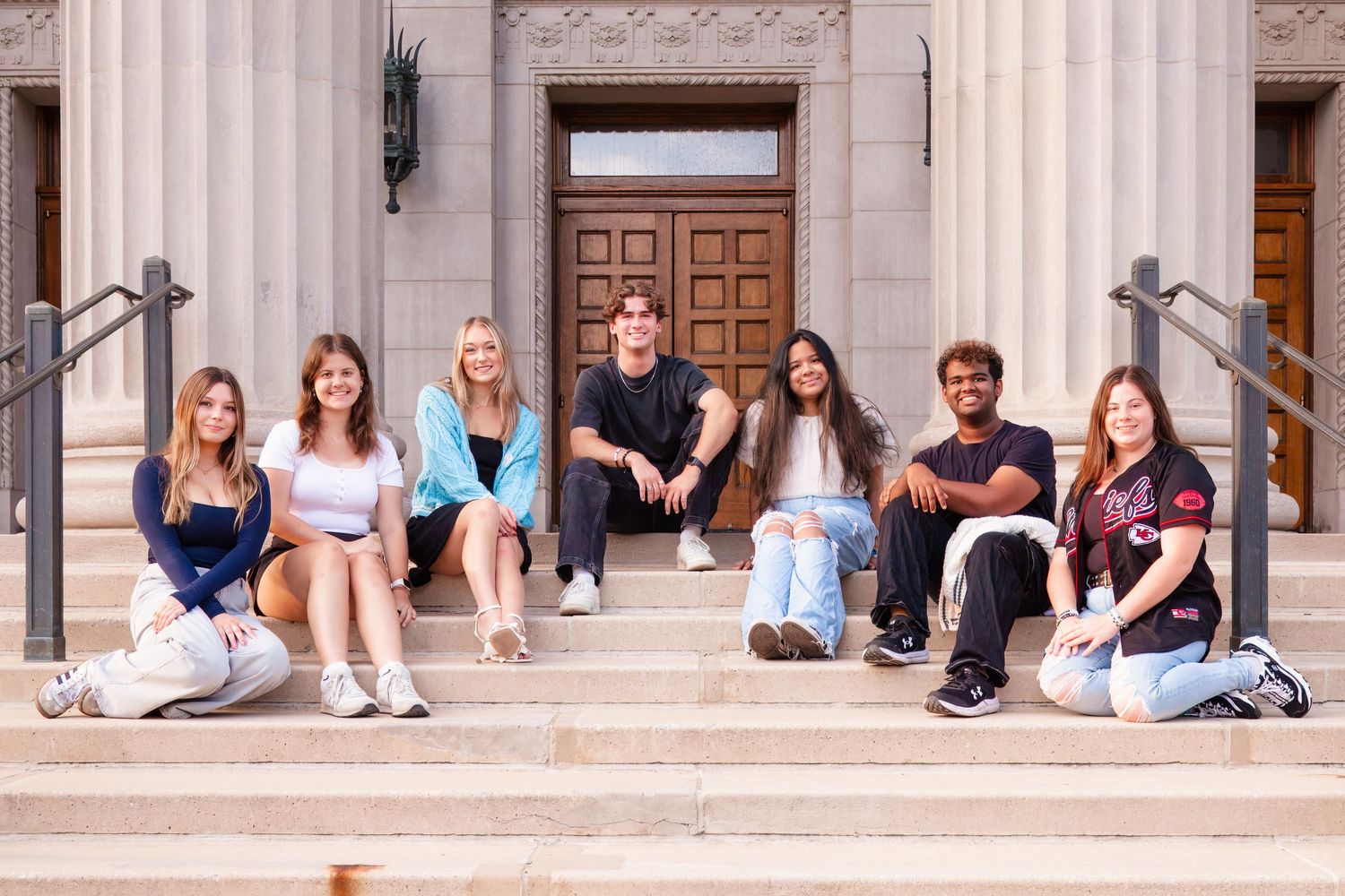 Seven college students smiling and sitting on stone steps outside a classic university building with large wooden doors.
