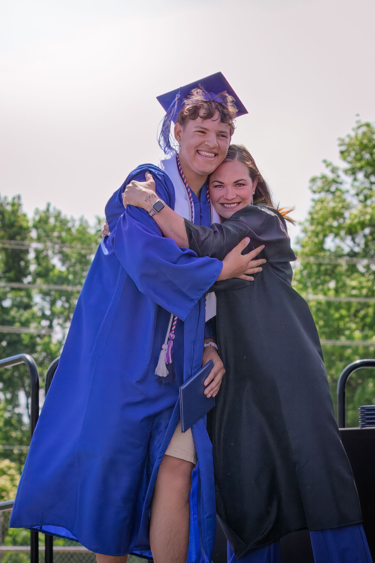 A graduate in blue cap and gown shares a warm embrace during commencement ceremonies on an outdoor stage.