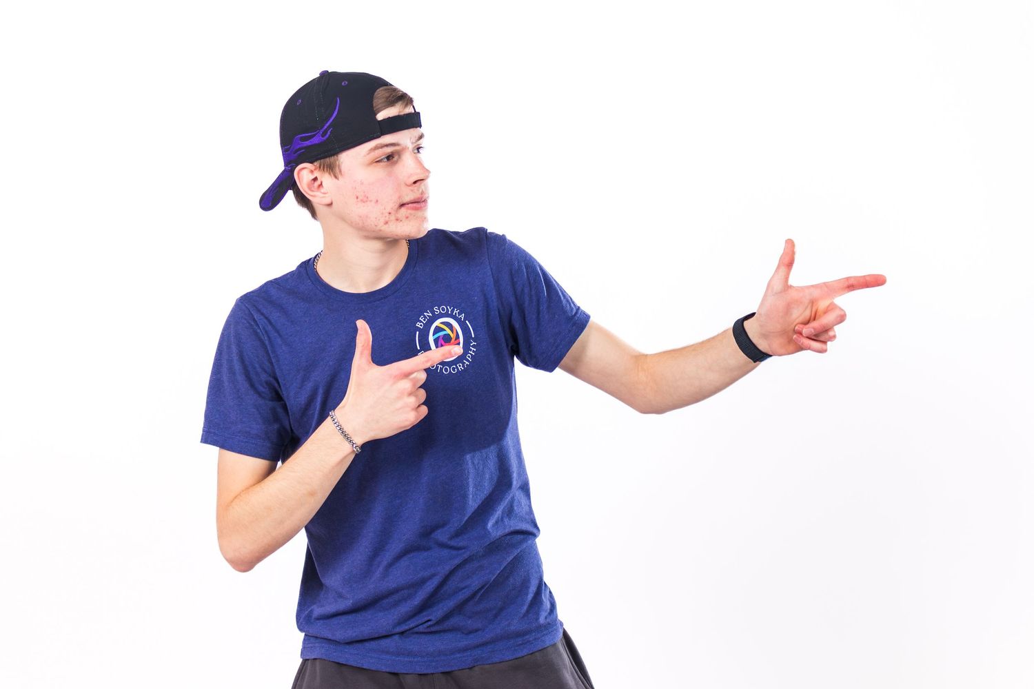 Young man in blue graphic t-shirt and backwards cap pointing finger guns against white background.