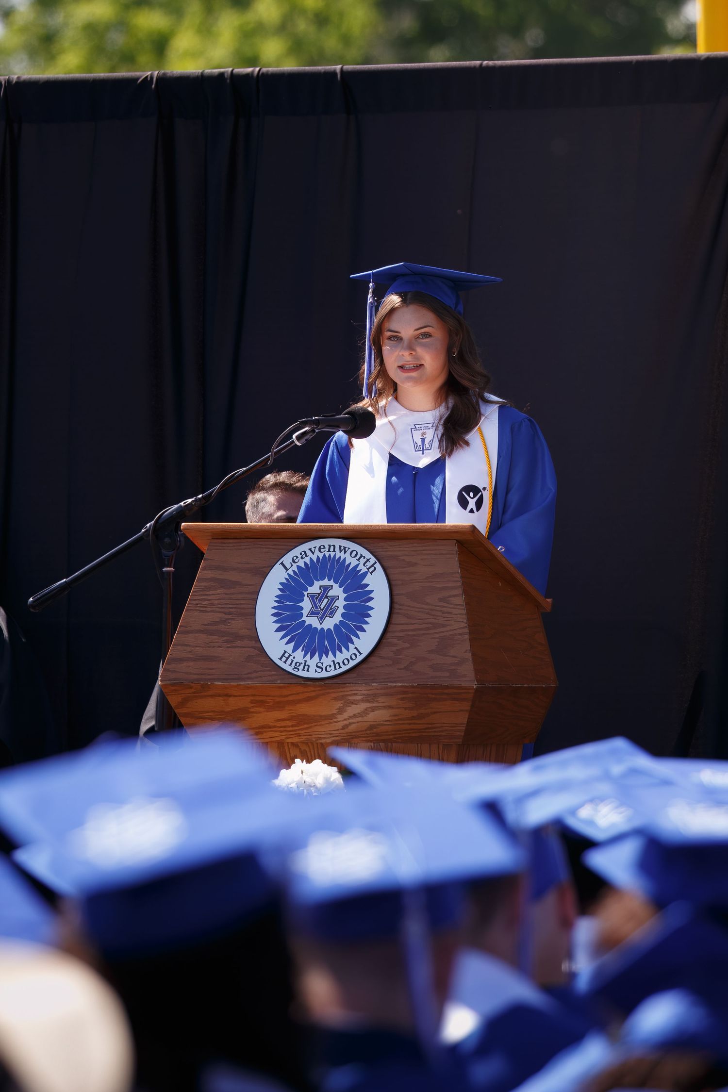 Student in blue graduation cap and gown delivers speech at podium during commencement ceremony.