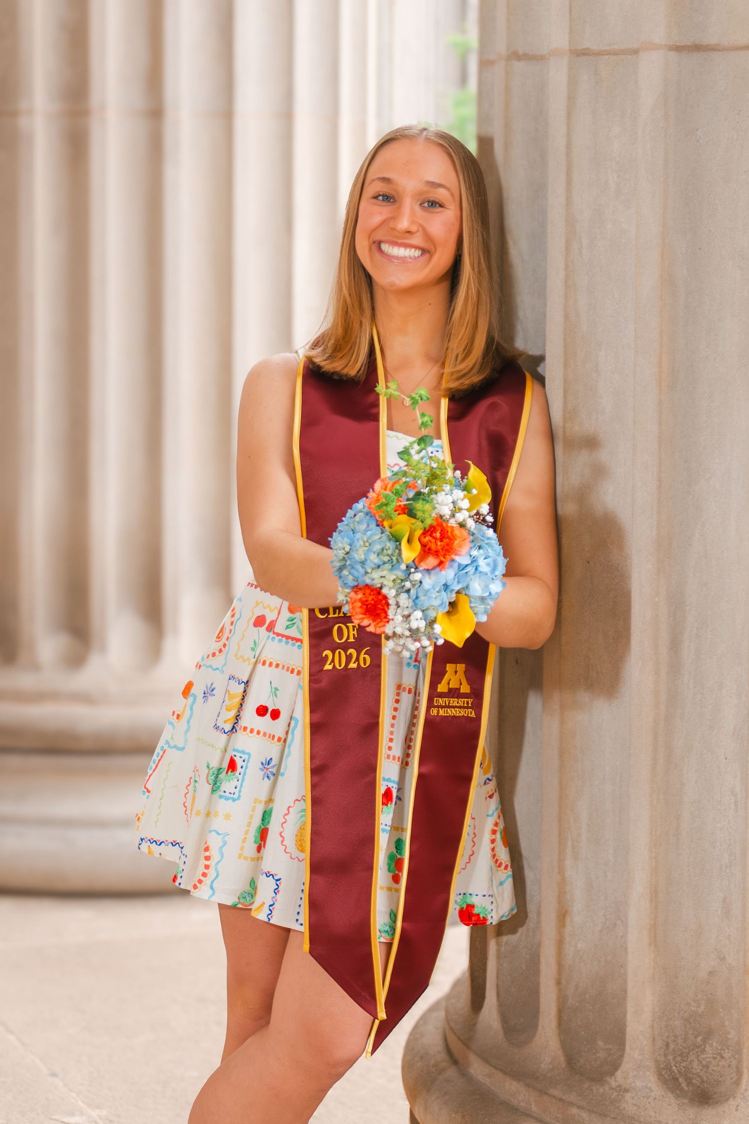 Smiling college student in University of Minnesota Class of 2026 graduation stole holding colorful bouquet by stone columns.