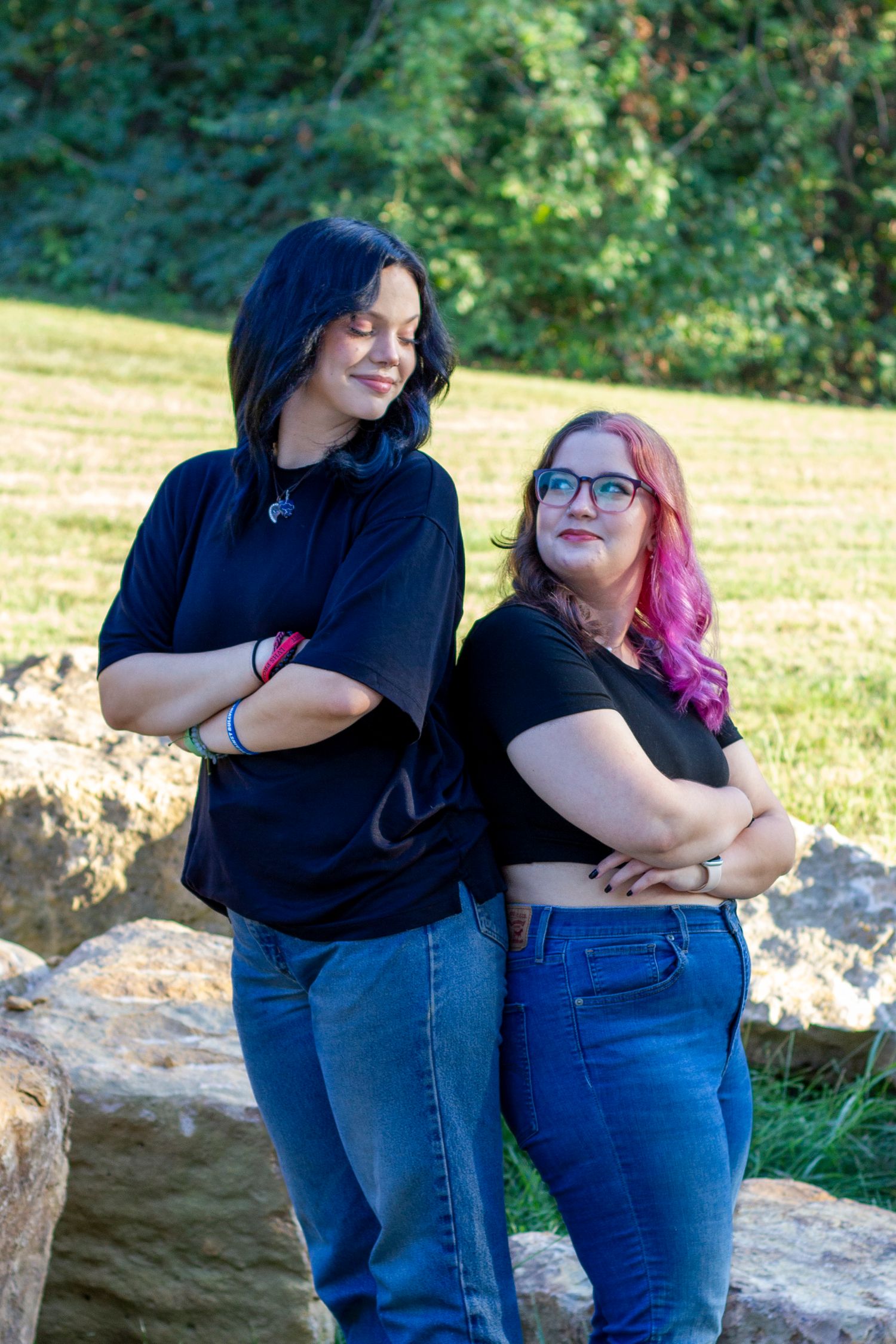 Two people with colorful hair wearing black tops and jeans pose casually on rocks outdoors on a sunny day.
