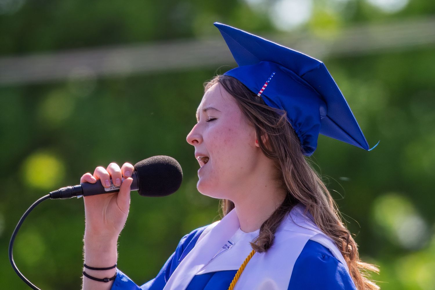 Graduate in bright blue cap and gown sings passionately into microphone during outdoor ceremony.