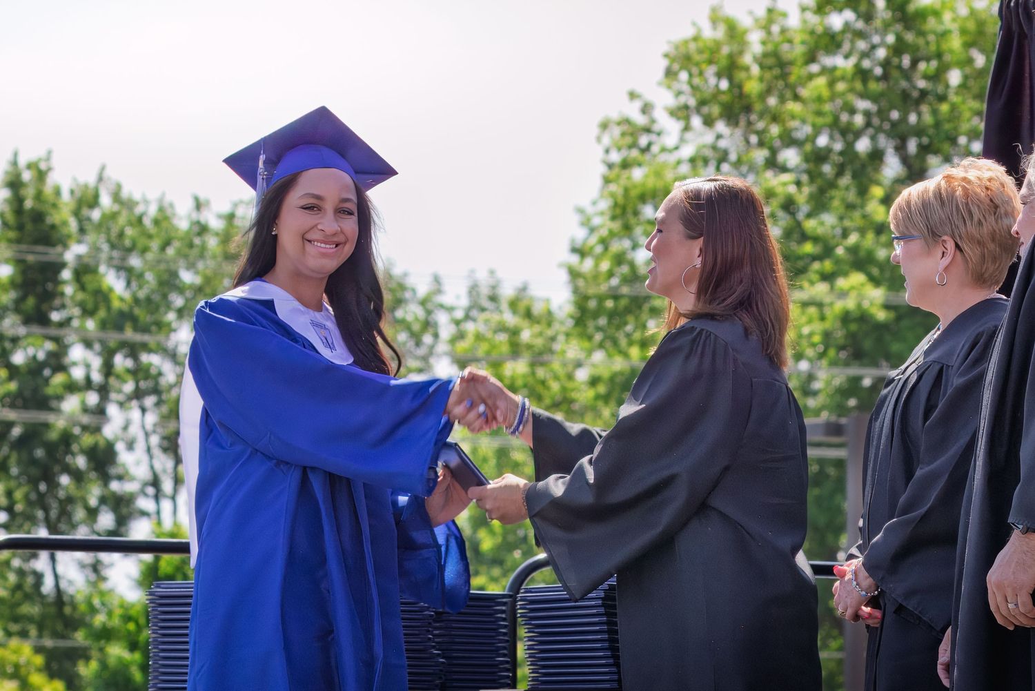 Graduate student in blue cap and gown receiving diploma during outdoor commencement ceremony.