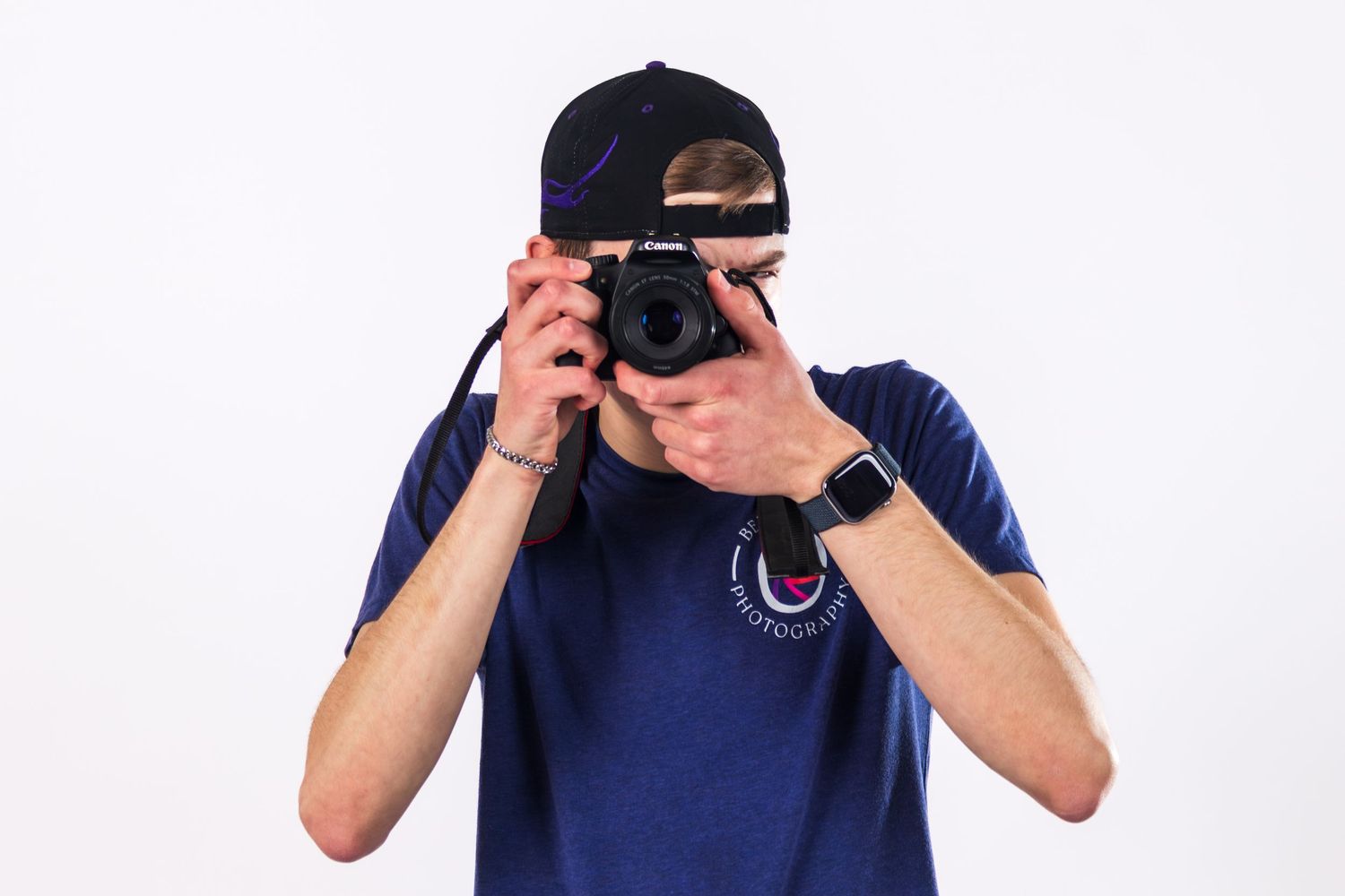 Man in blue t-shirt and black cap photographing with a DSLR camera against a white background.