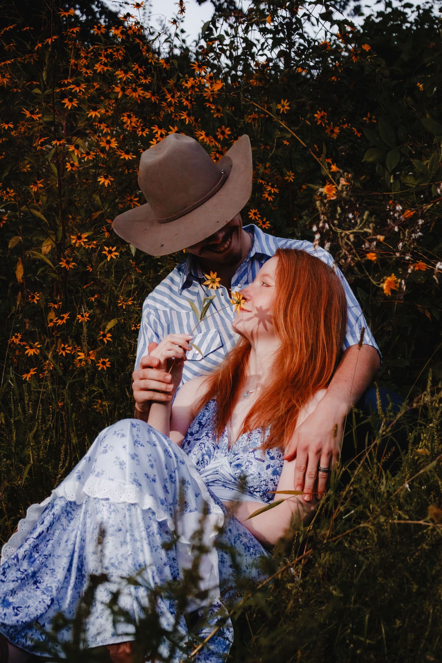 A romantic couple in a blue dress and brown hat embrace on the grass during sunset with autumn foliage in the background.