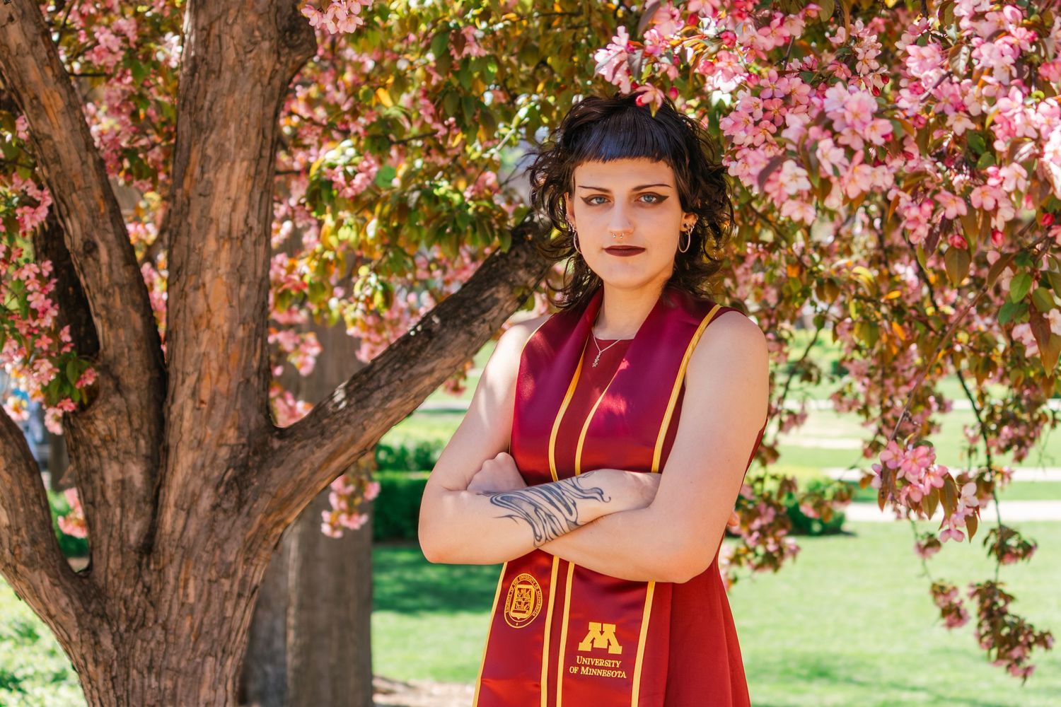 Graduate in maroon cap and gown poses confidently with crossed arms before blooming pink cherry blossom tree outdoors.