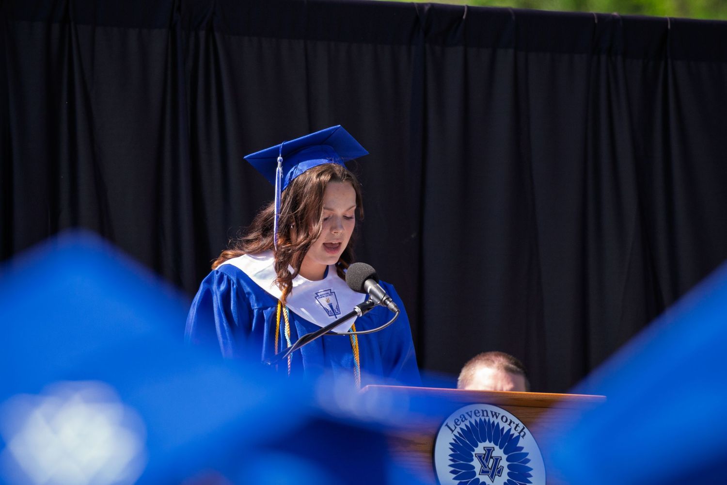 A graduate in blue cap and gown stands at a podium during an outdoor commencement ceremony.