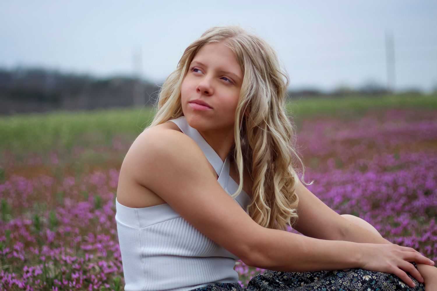 Person in white tank top sitting in vibrant purple wildflower field on cloudy summer day.