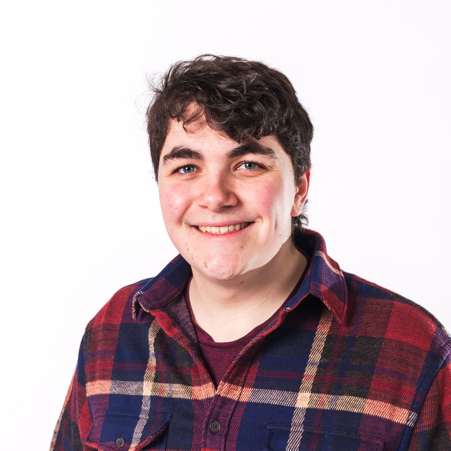 Young person smiling, wearing a red plaid flannel shirt, with dark curly hair against a white background.