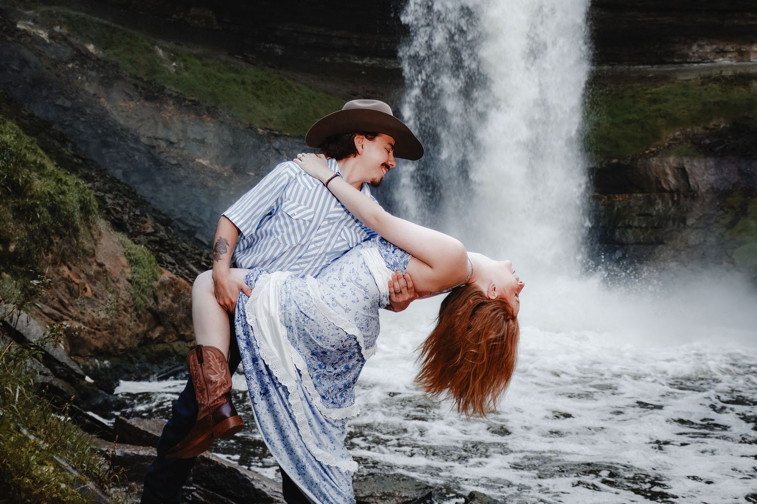 A couple in flowy dresses poses romantically near a cascading waterfall in a scenic mountain landscape.