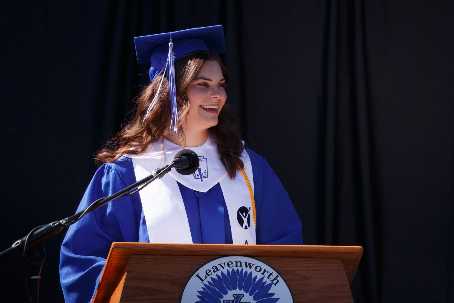 A graduate delivering a speech at a podium wearing a blue cap and gown.
