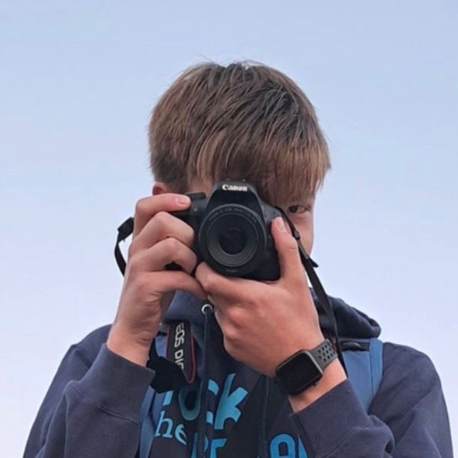 A photographer wearing a dark hoodie holds up a Canon DSLR camera against a light blue sky background.