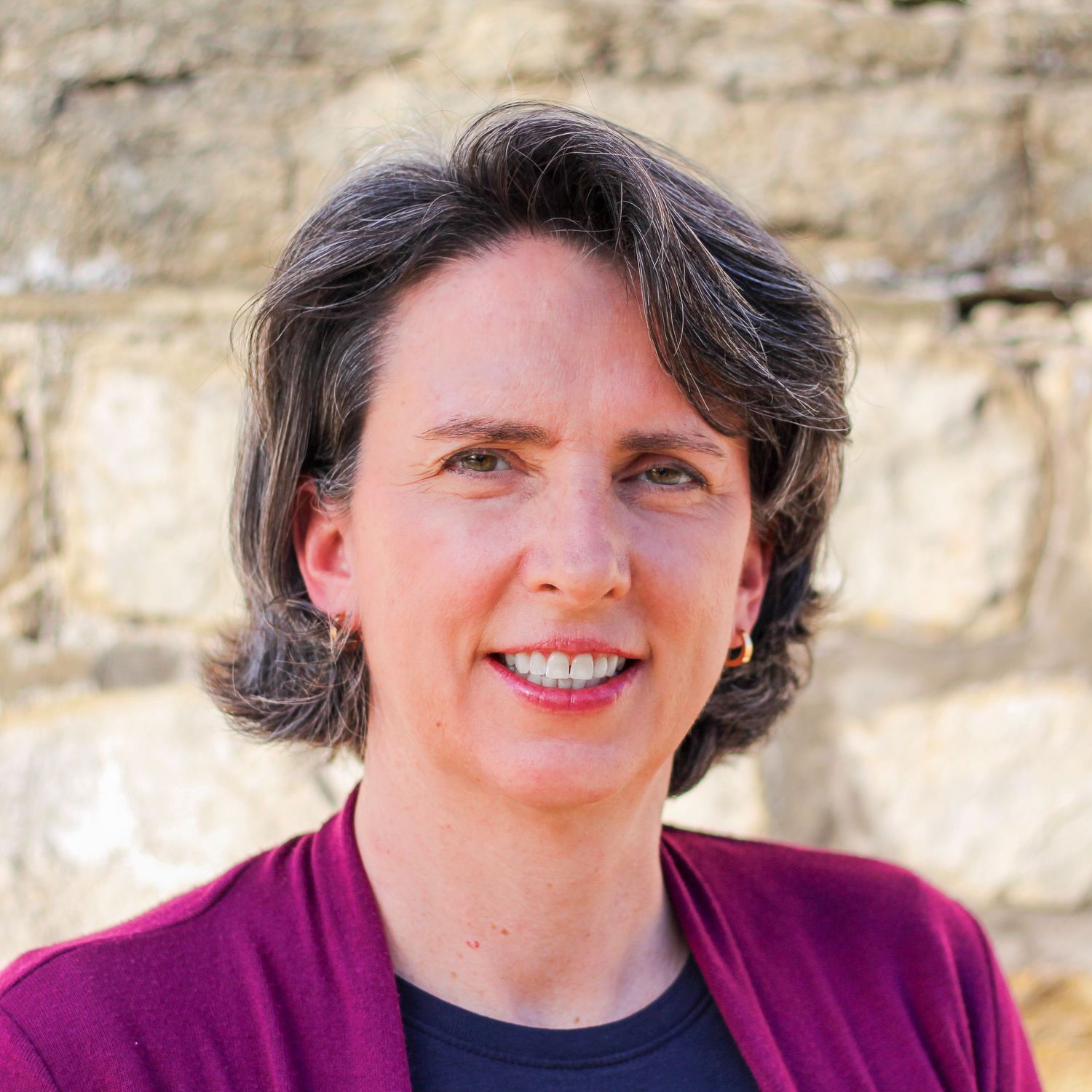 Professional headshot portrait of someone in a magenta sweater and black shirt against a stone or concrete wall background.