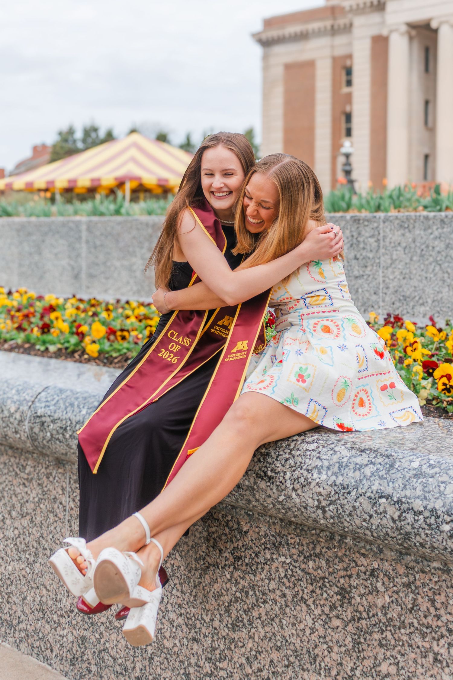 Two young women in graduation stoles share a warm hug while sitting on a stone ledge surrounded by colorful flowers.