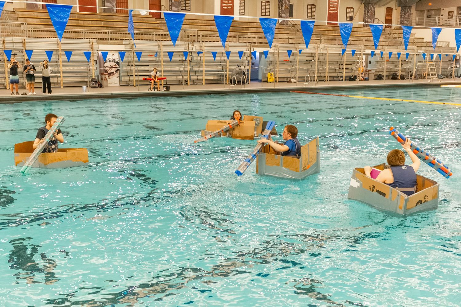 Children racing homemade cardboard boats in an indoor swimming pool during a fun engineering challenge event.