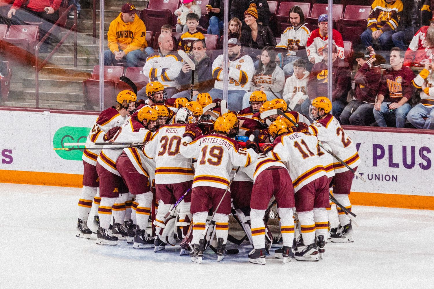 Hockey team huddles together on ice during game while fans watch from behind glass at indoor arena.