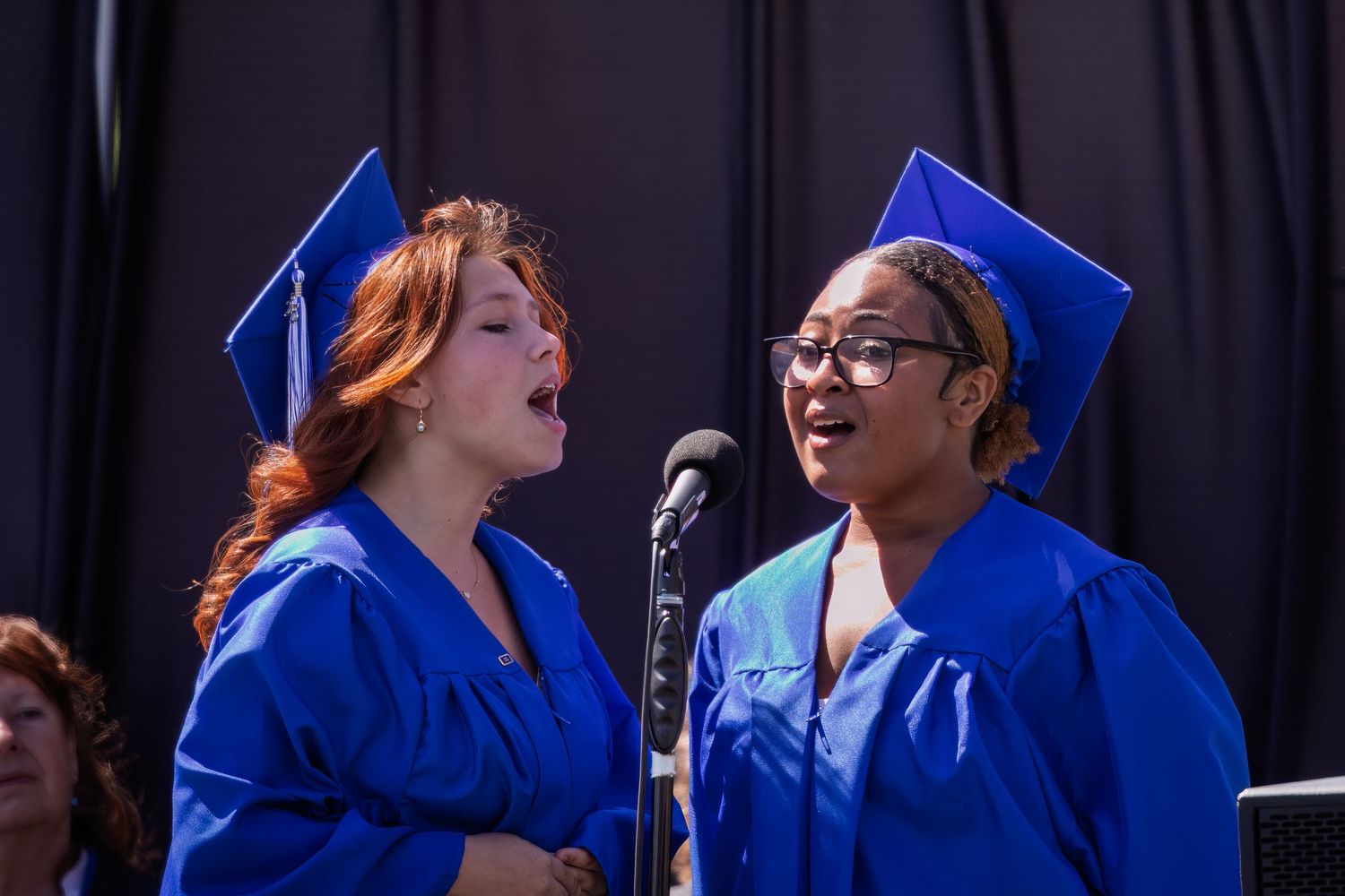 A row of graduates in blue caps and gowns smile and celebrate during a commencement ceremony.