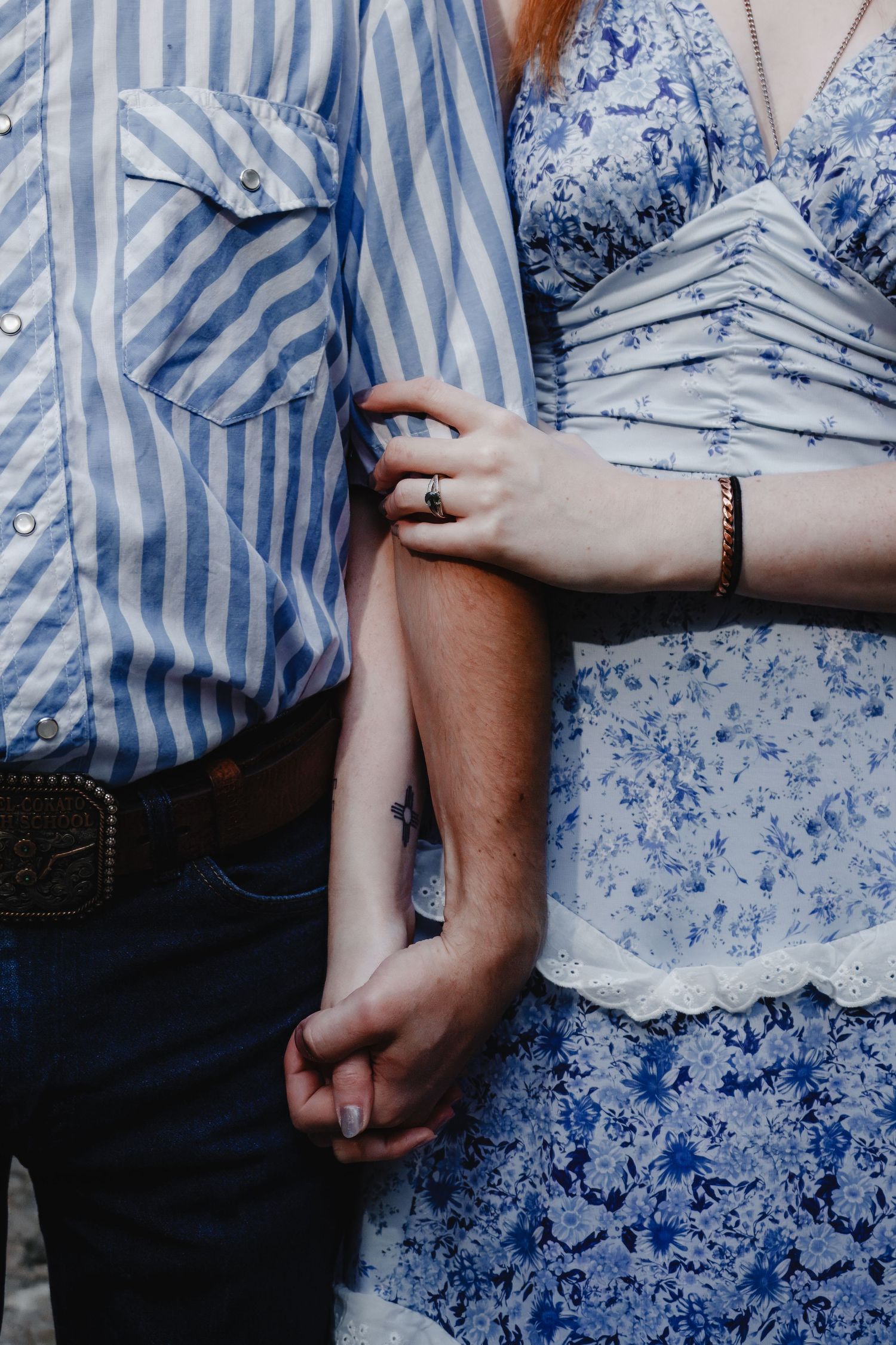 Close-up of couple holding hands, wearing blue striped shirt and floral blue dress with jewelry details.