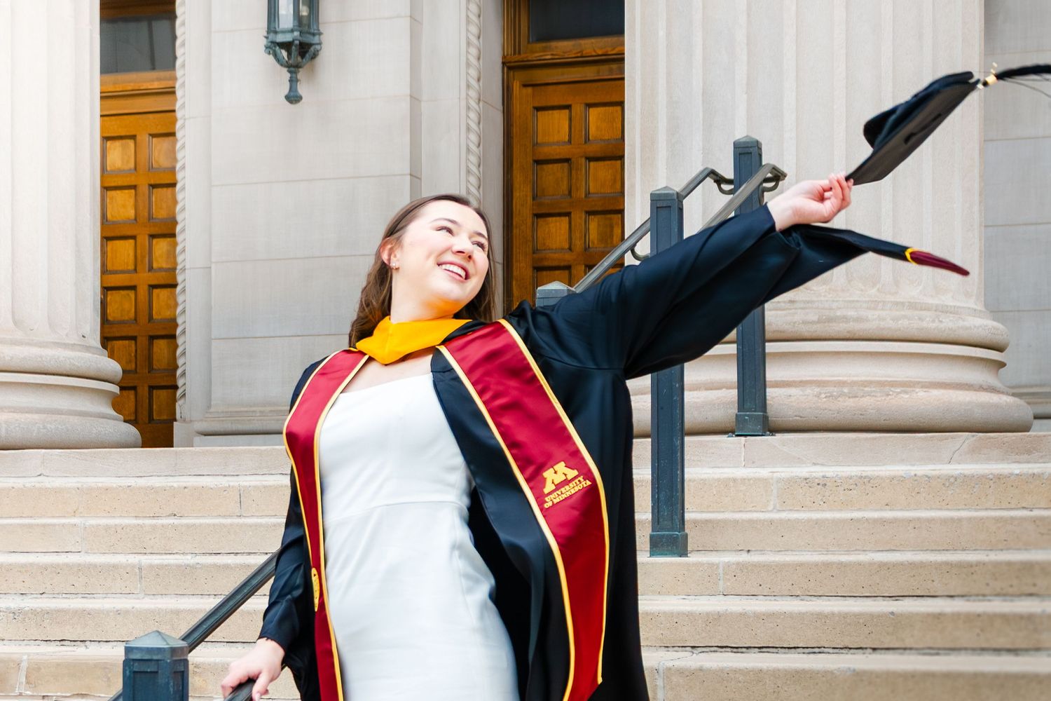 Woman in graduation cap and gown celebrating on university steps, tossing cap in the air with joy.