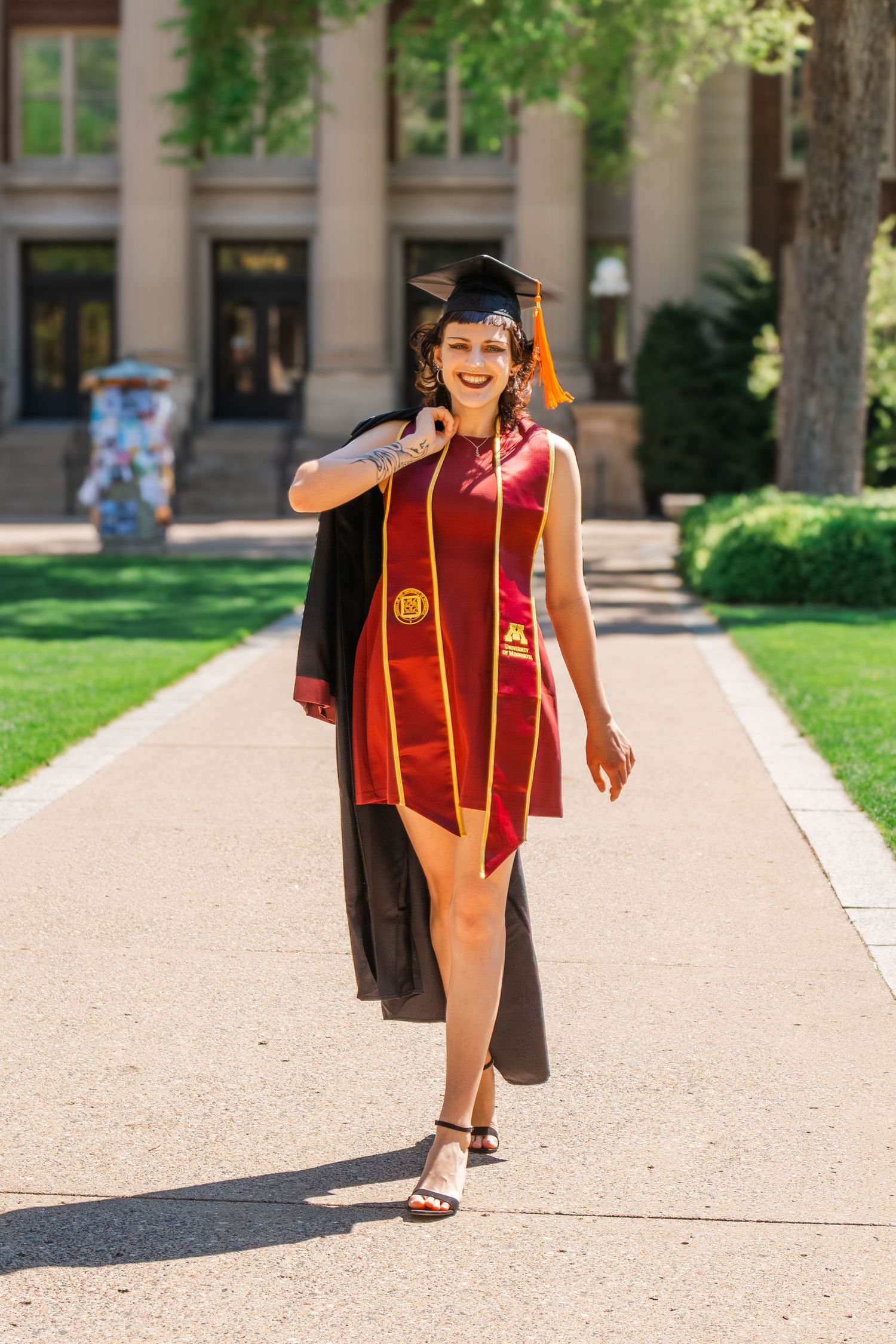 Smiling graduate in red dress, cap, and gown walks outdoors on campus pathway on a sunny day.