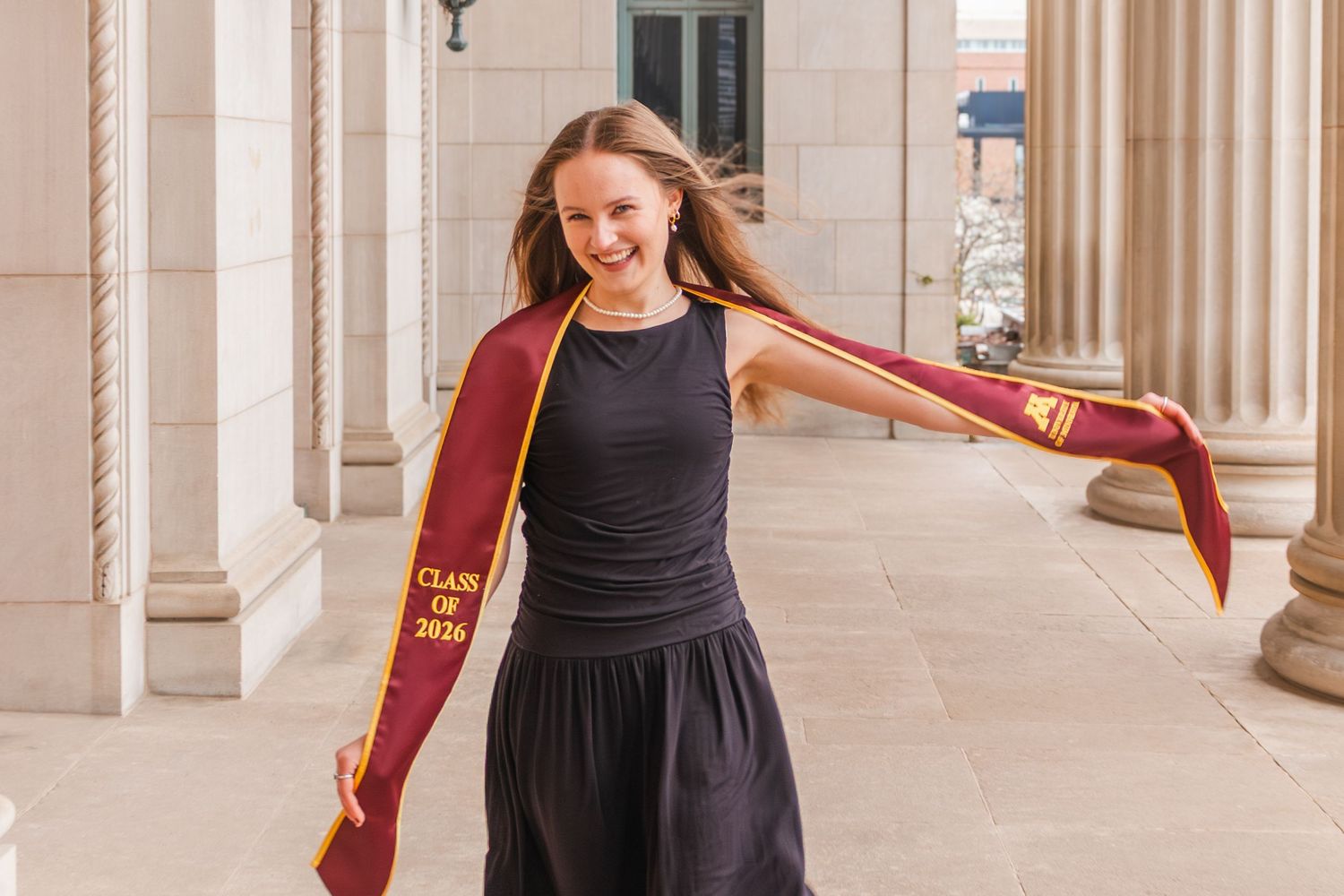 Smiling graduate twirling a maroon Class of 2020 graduation stole between stone columns.