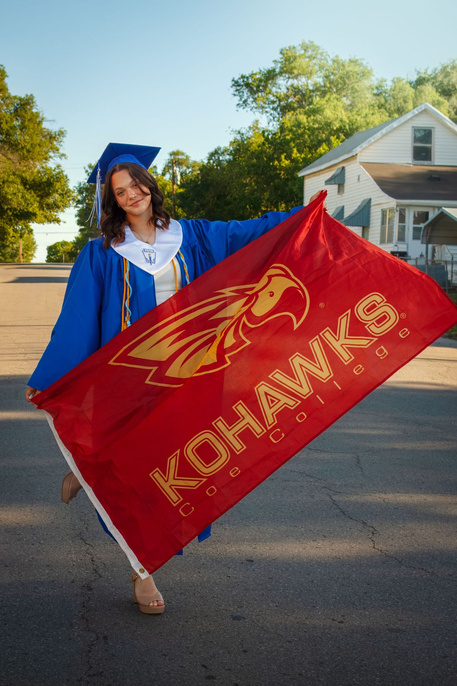 Graduate in blue cap and gown holds a large red Kohawks flag outside on a sunny day.