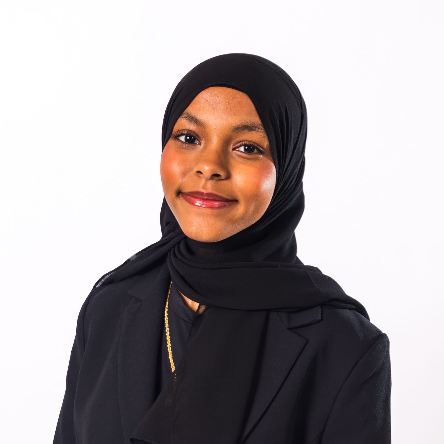 Young woman wearing a black hijab and black blazer with gold necklace, smiling against white background.