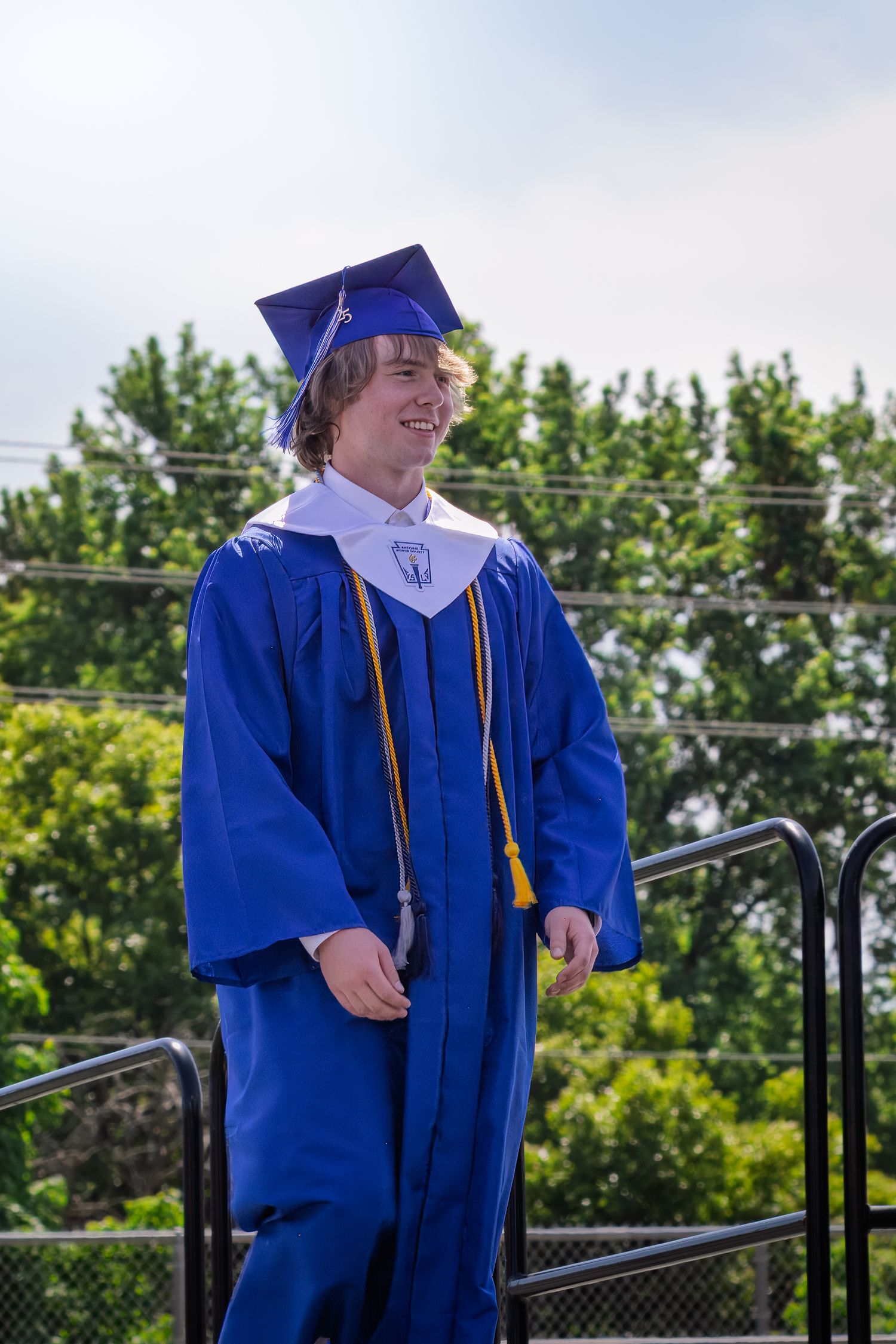 A graduate wearing blue academic regalia and honors cords stands smiling on stage during graduation day.