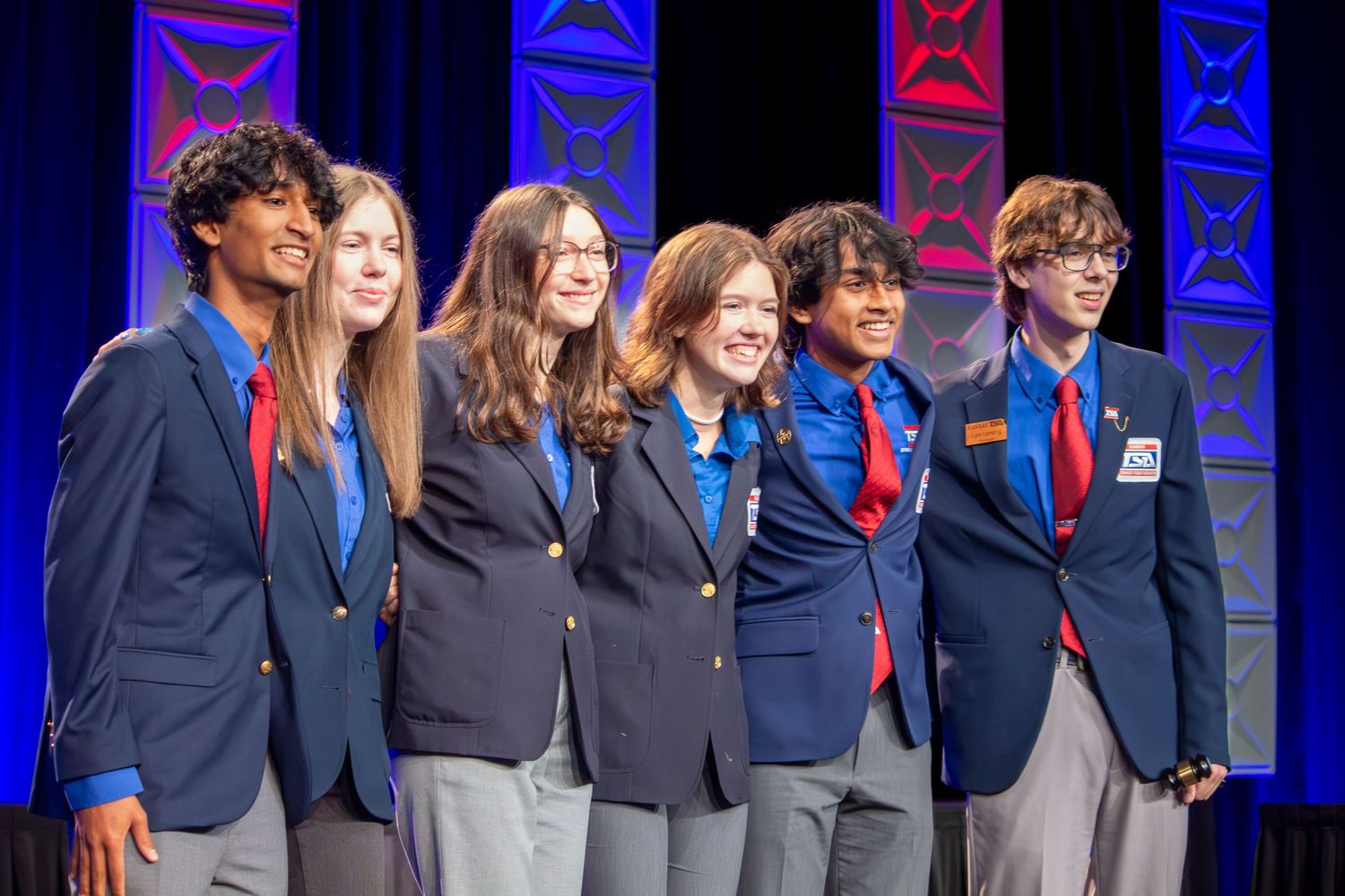 Students in navy blazers and red ties line up on stage during a formal academic event or ceremony.