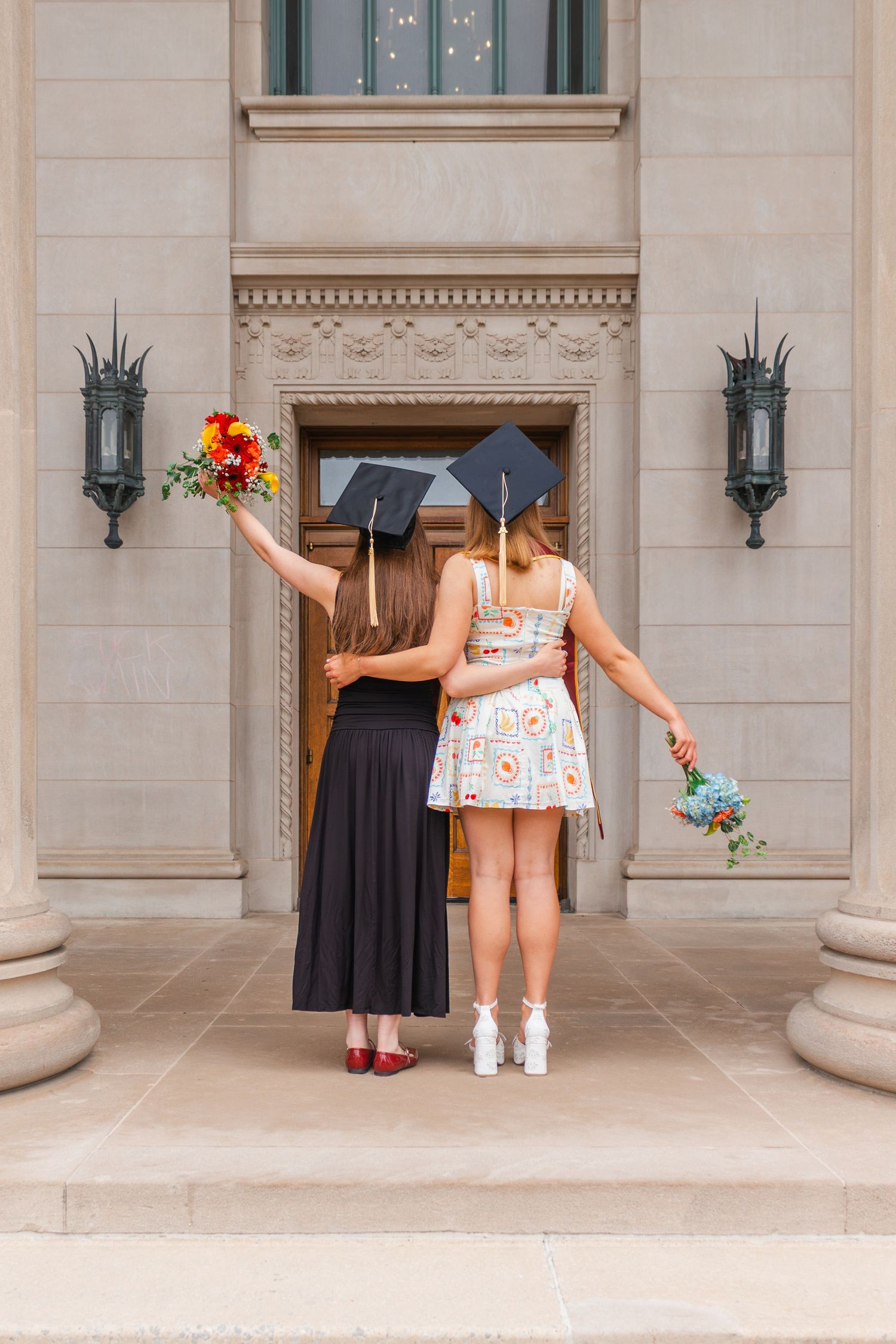 Two graduates in black gowns and caps hold flower bouquets while embracing on university building steps.