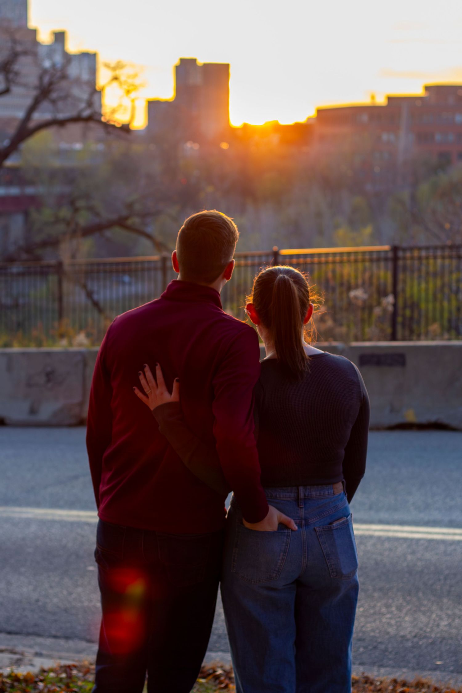 A couple watches the sunset over city buildings from a rooftop viewpoint on an autumn evening.