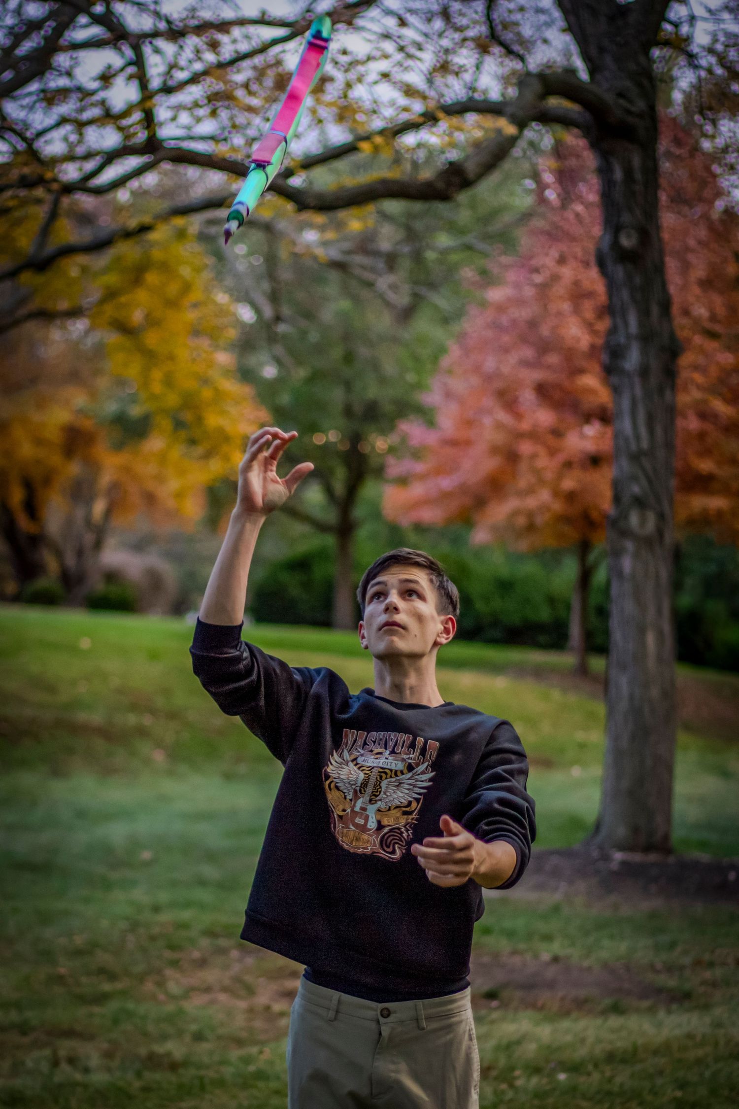 Someone wearing a dark sweater tosses a colorful toy in the air at a park with autumn foliage in the background.