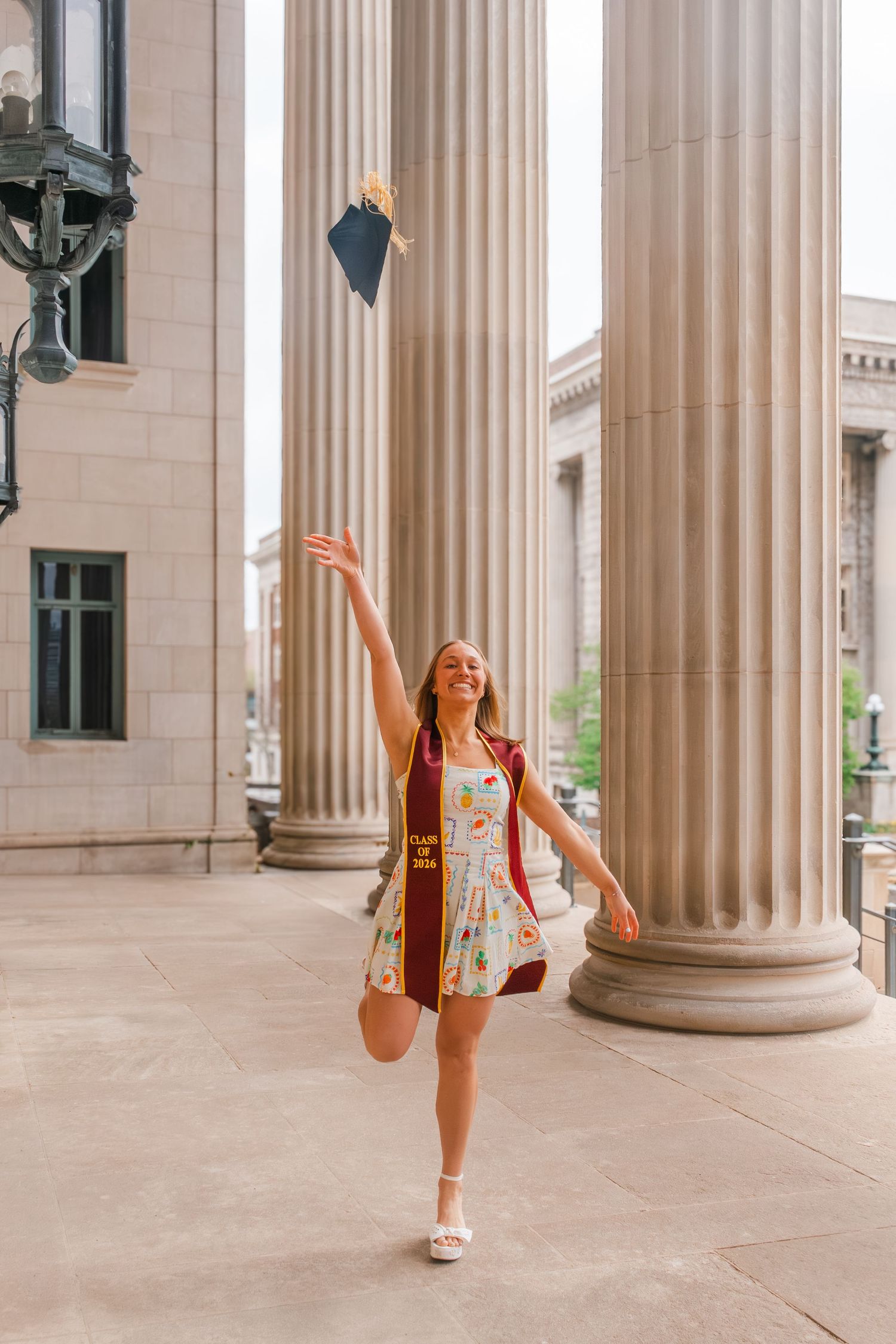 A joyful graduate tosses her cap in the air while balancing on one leg beside grand stone columns.