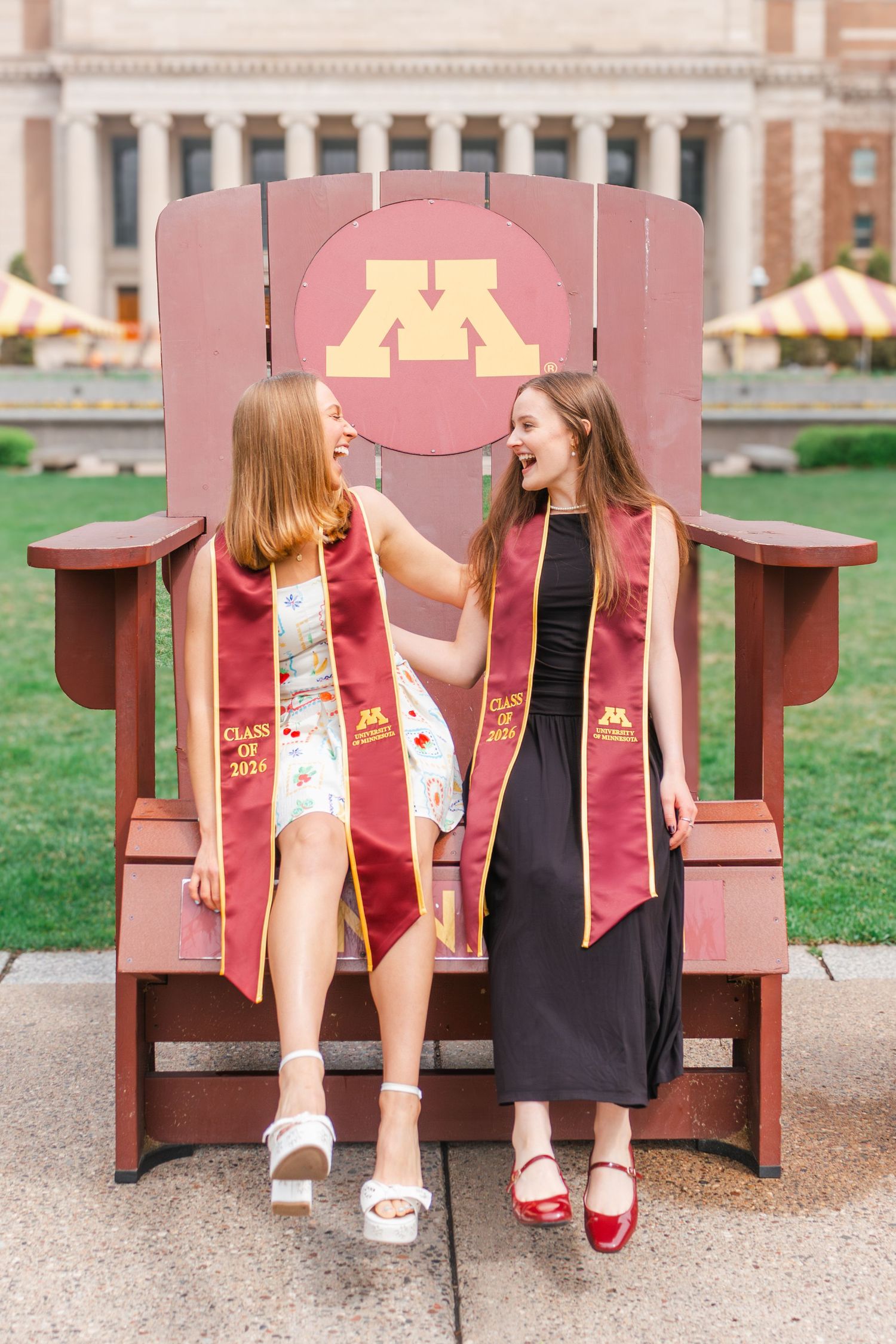 Two female University of Minnesota graduates wearing maroon stoles sit on a giant Adirondack chair on campus.