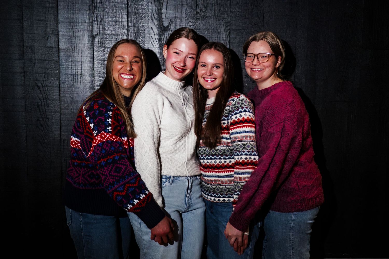 Four people wearing cozy winter sweaters with festive patterns pose together against a dark background.