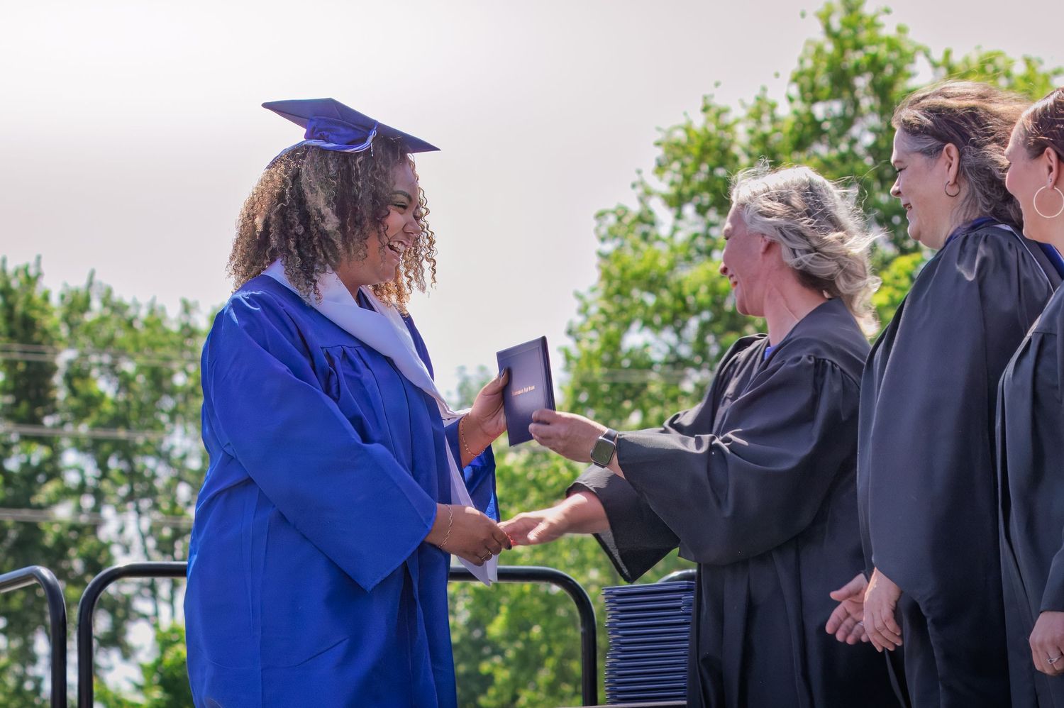 Graduate in blue cap and gown receives diploma during outdoor commencement ceremony.