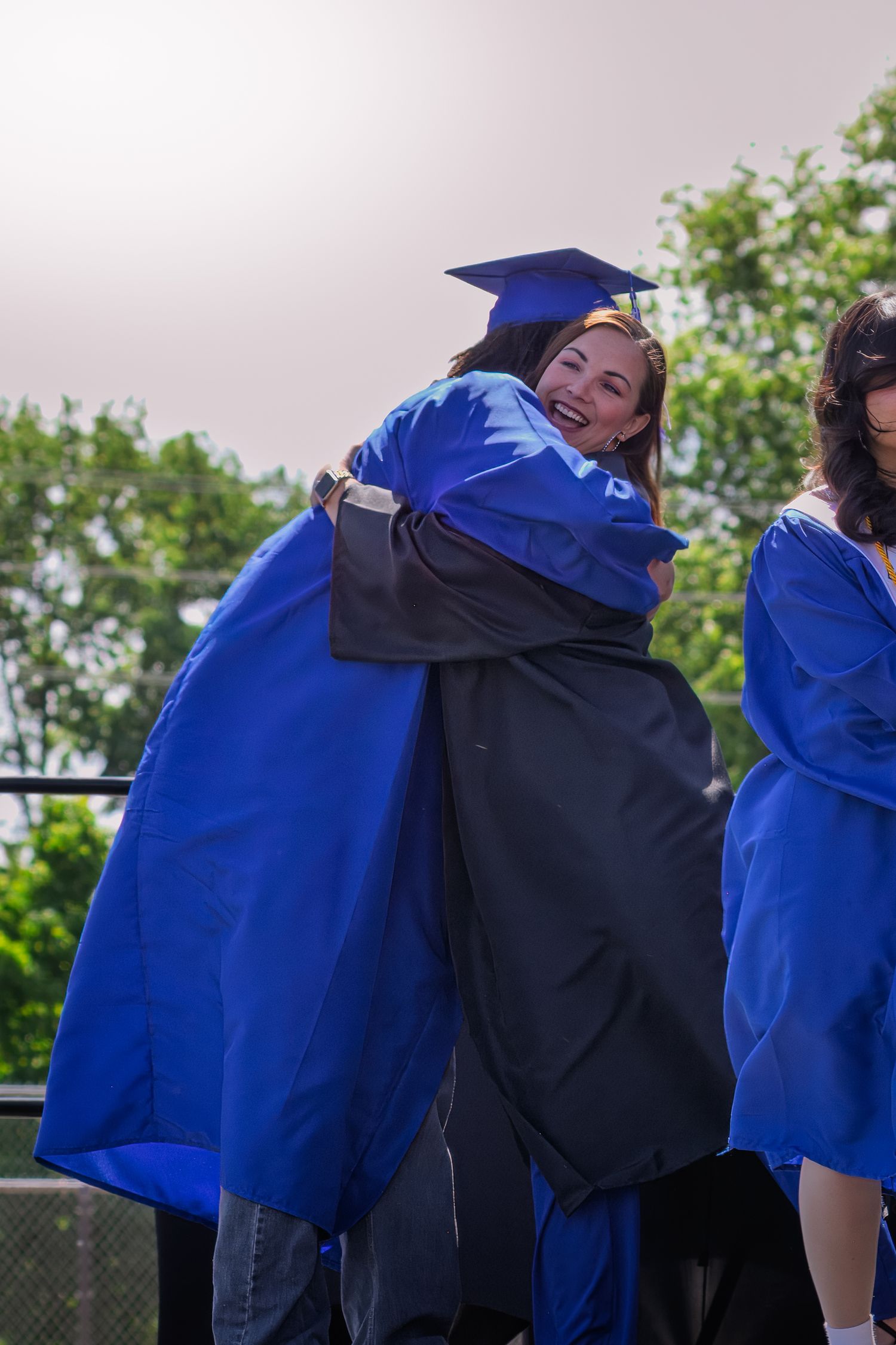 Two graduates in bright blue commencement robes share a celebratory hug during graduation ceremony outdoors.