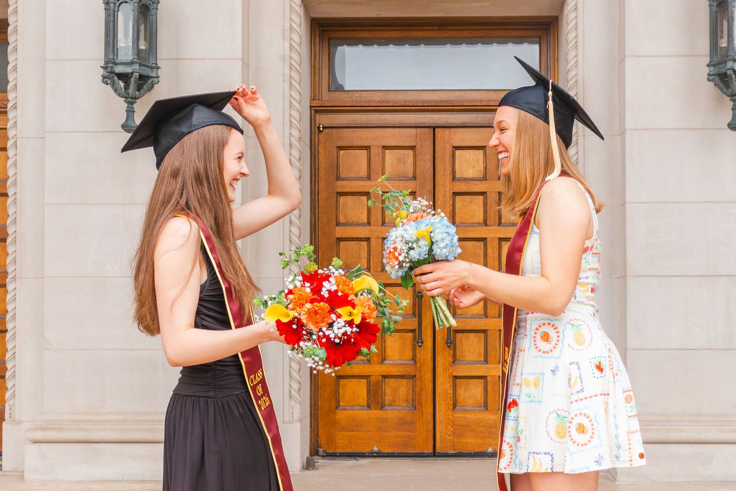 Two female graduates in caps holding colorful bouquets, celebrating outdoors near a wooden door.