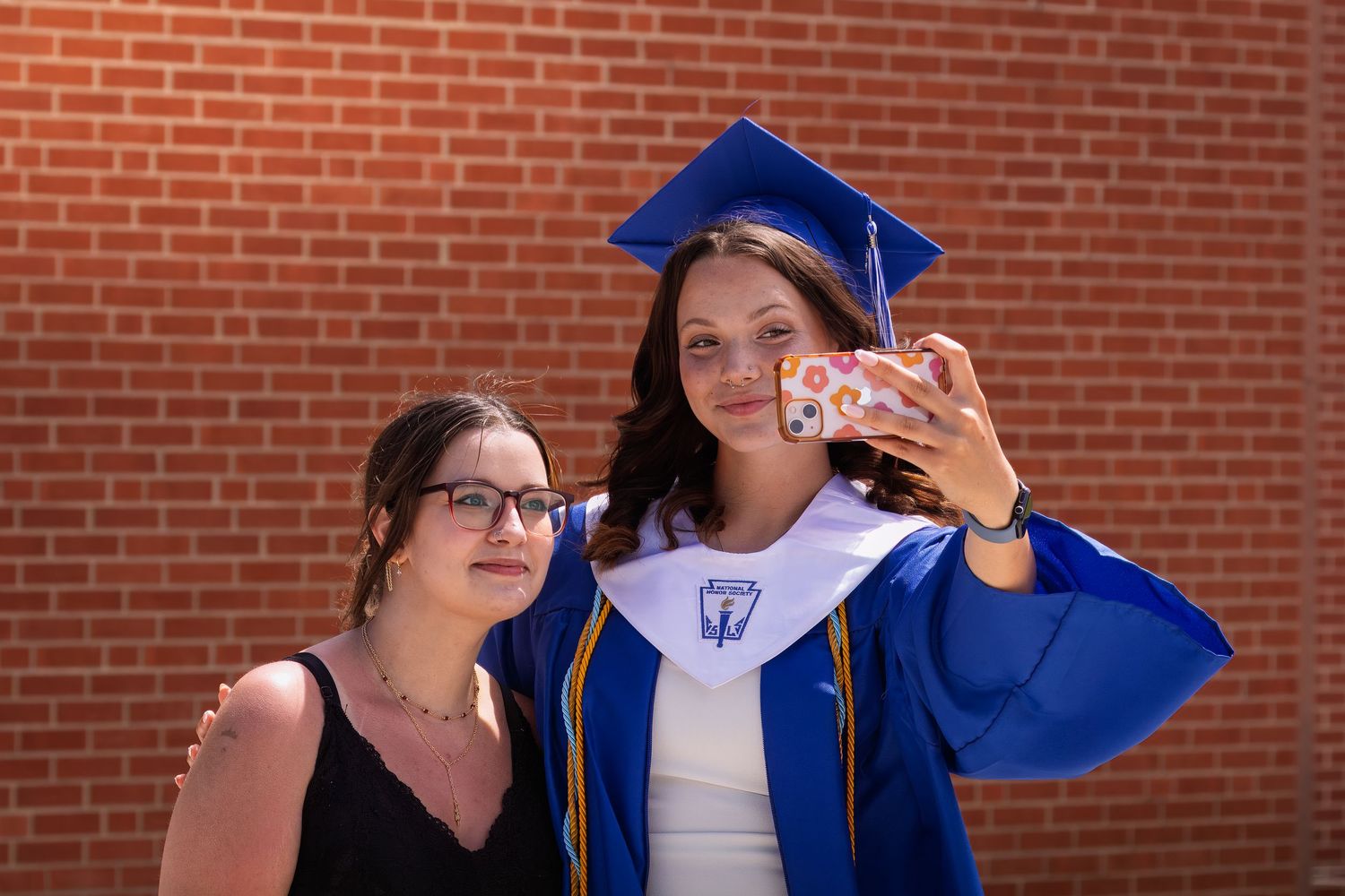 Two people pose together against a brick wall with one wearing a blue graduation cap and gown taking a selfie.