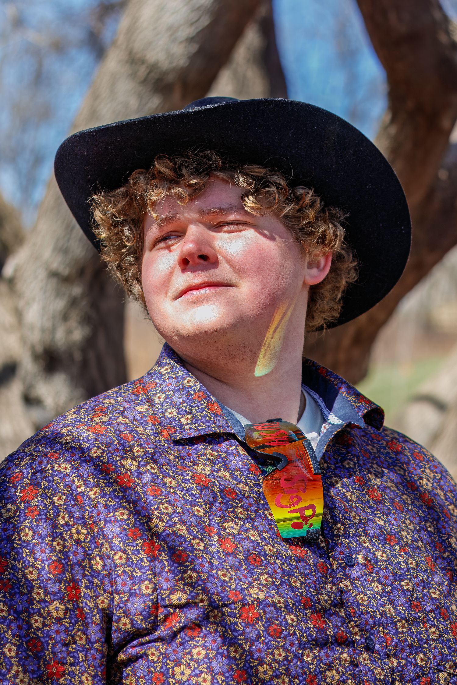 Person wearing a patterned shirt and black hat with rainbow sunglasses outdoors.