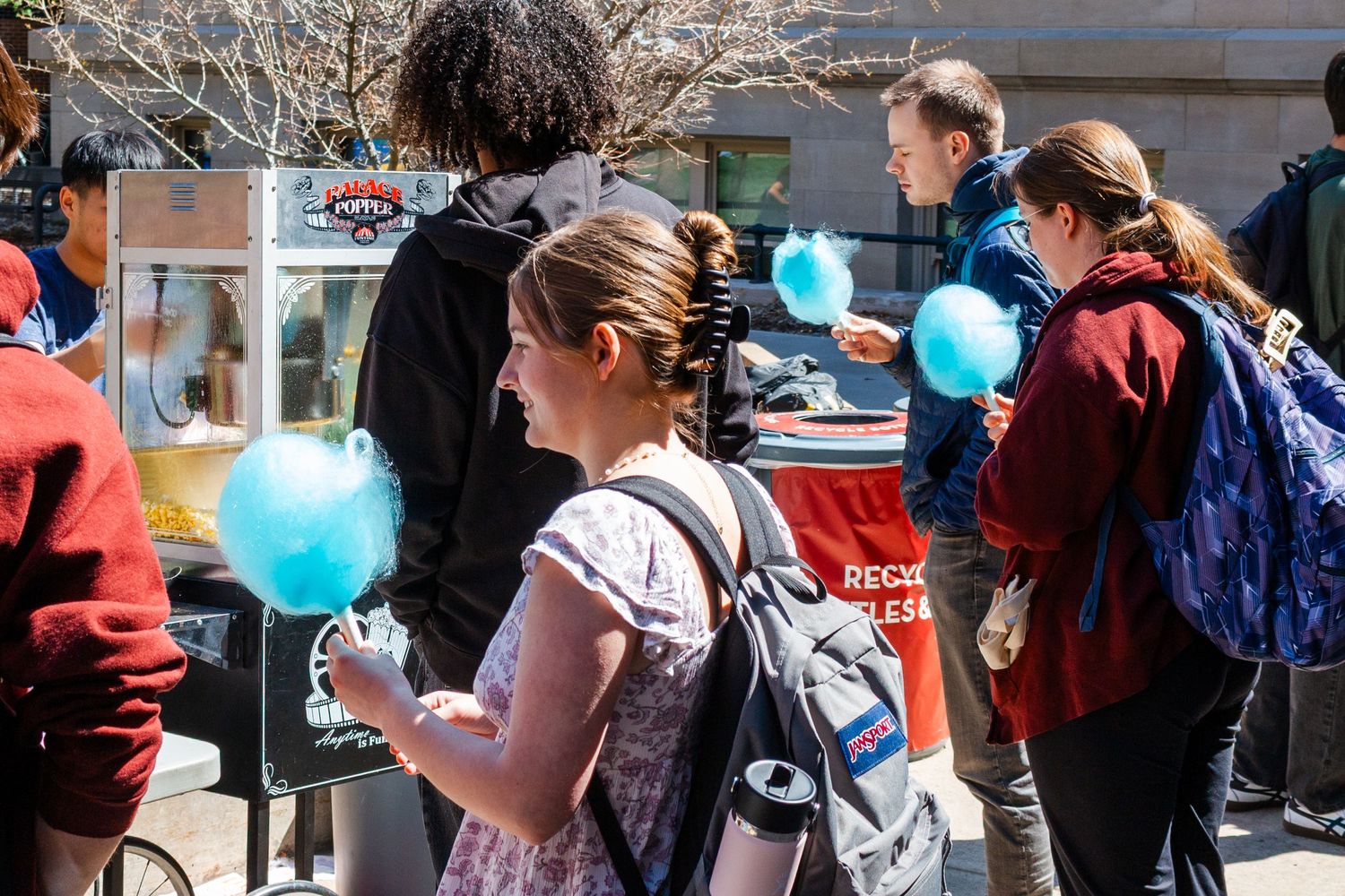 Students gather outdoors holding blue cotton candy at a sunny campus event with food vendors in the background.