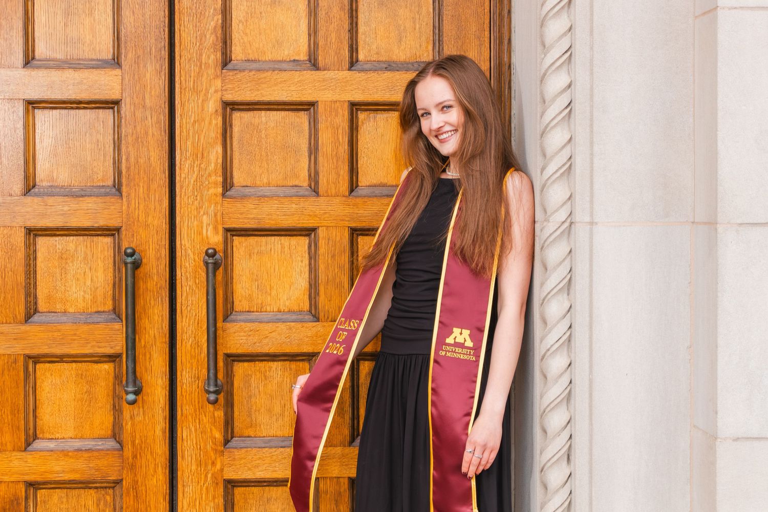 Young woman in black dress with maroon graduation stole smiling beside ornate wooden doors.