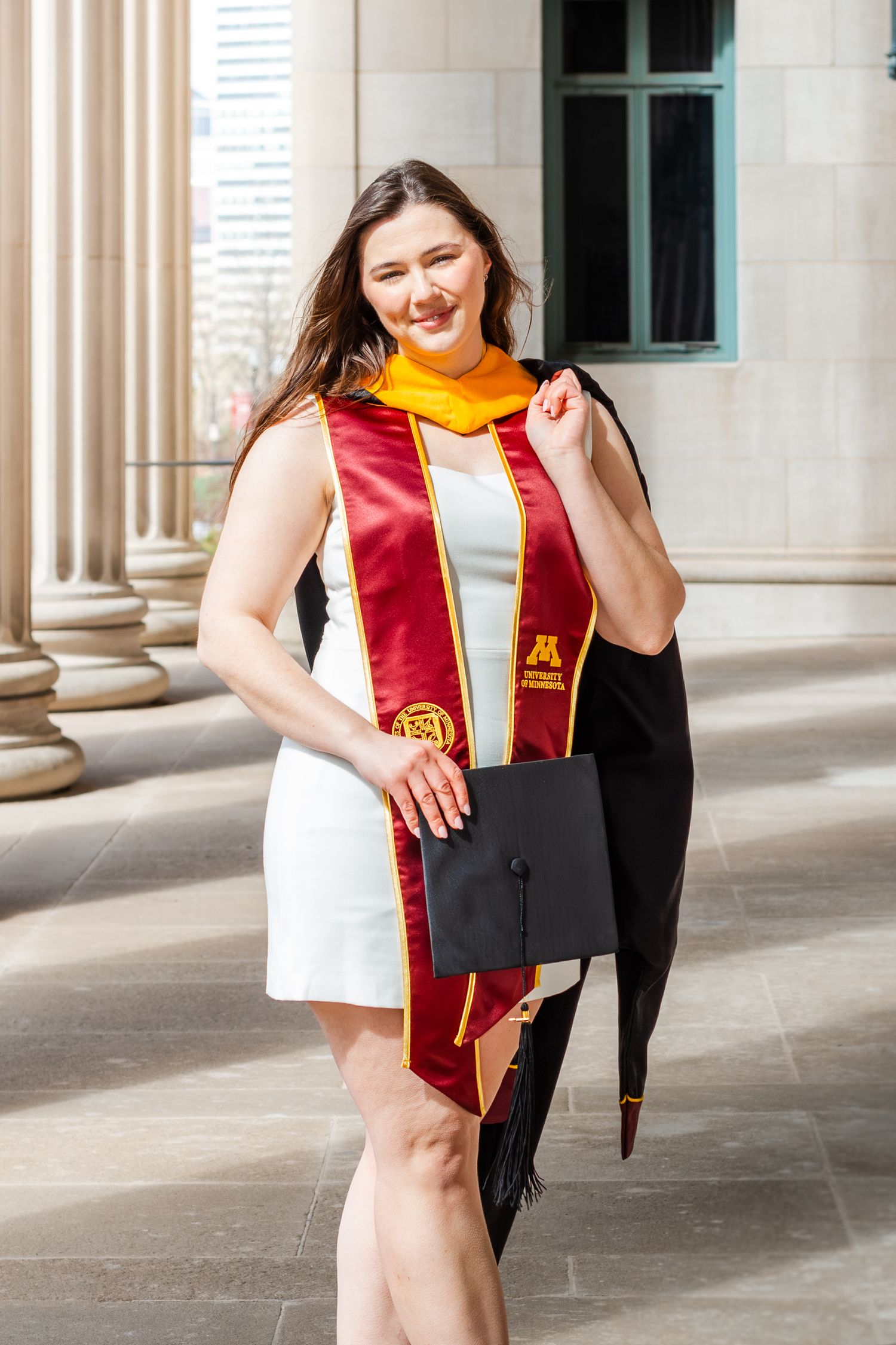 Young woman in white dress wearing University of Minnesota graduation stole, holding cap outdoors near stone pillars.