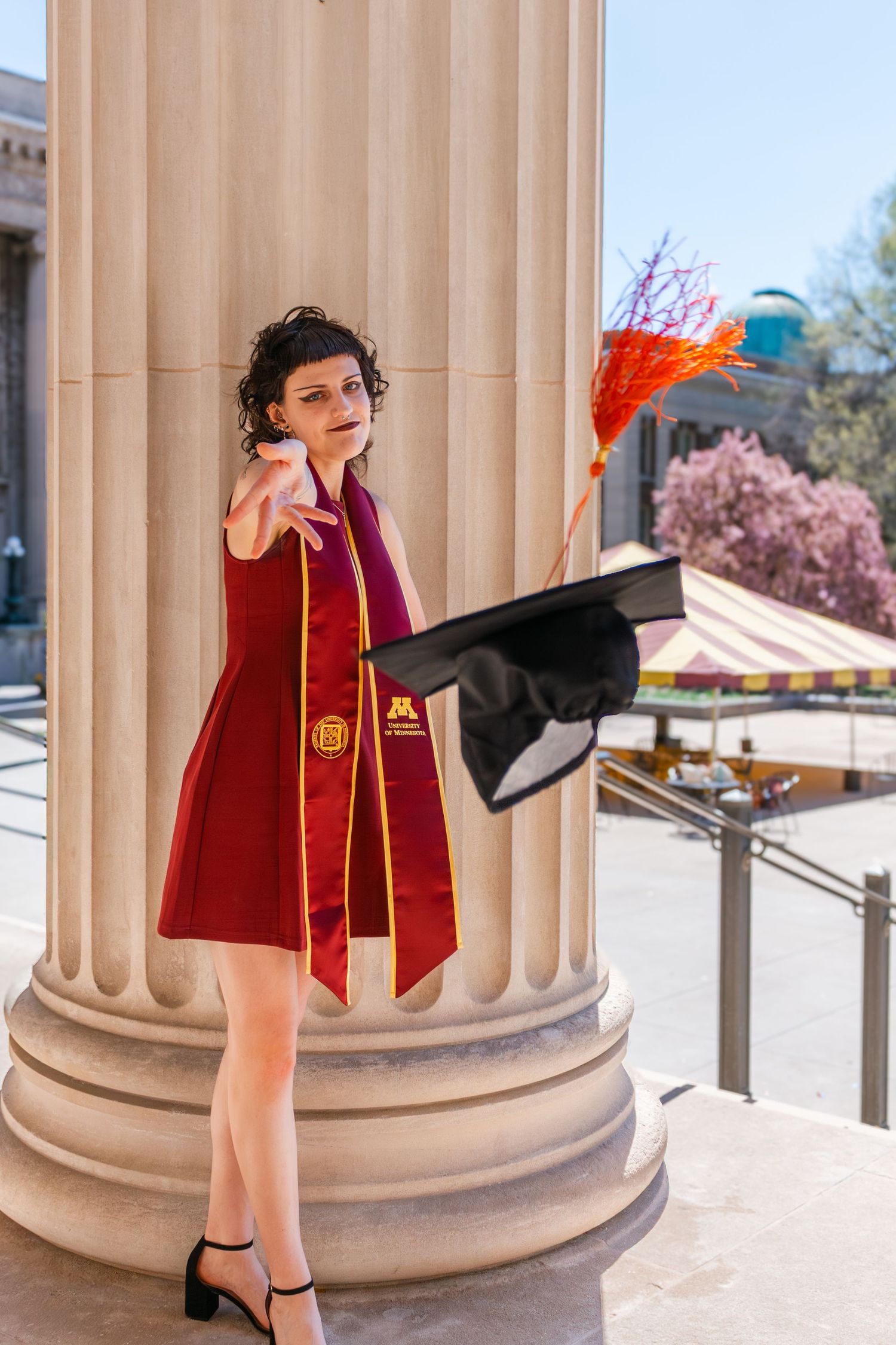 University of Minnesota graduate in red regalia tosses cap beside campus column on sunny day.