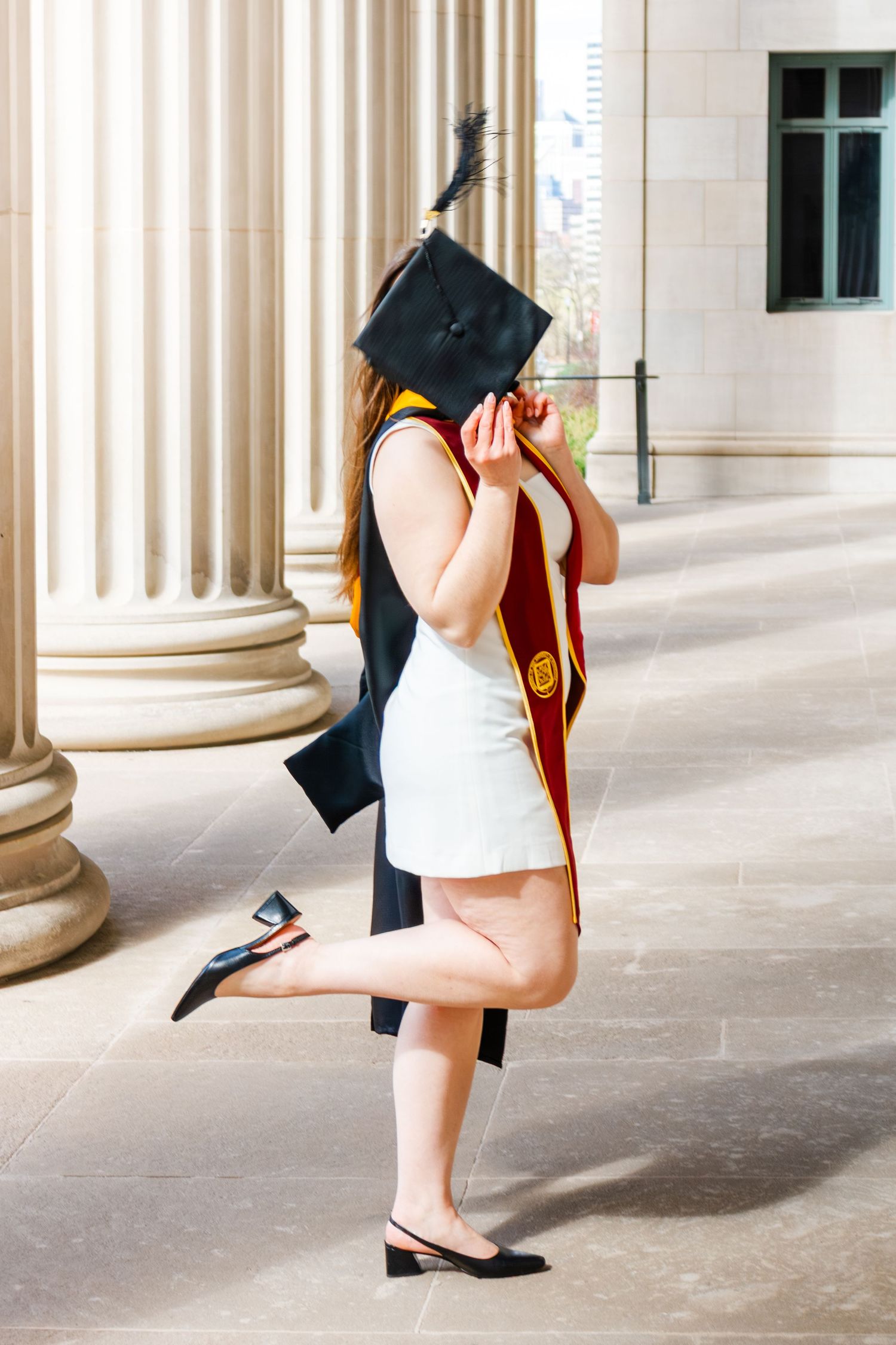 Graduate in cap and gown holding mortarboard over face, wearing white dress and maroon stole, posing by columns.