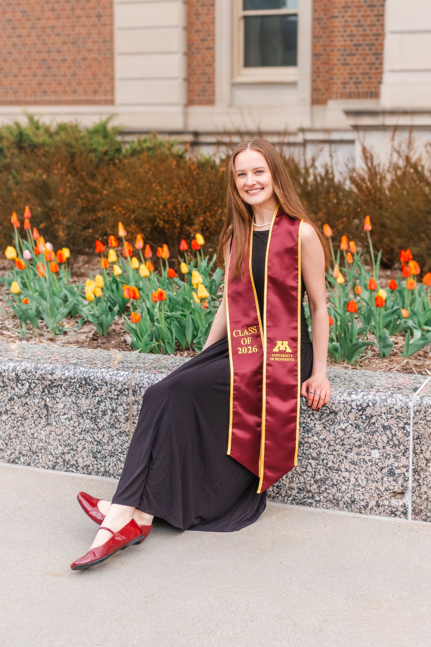 Smiling female graduate in maroon Class of 2026 stole poses in black gown beside colorful tulips on campus.